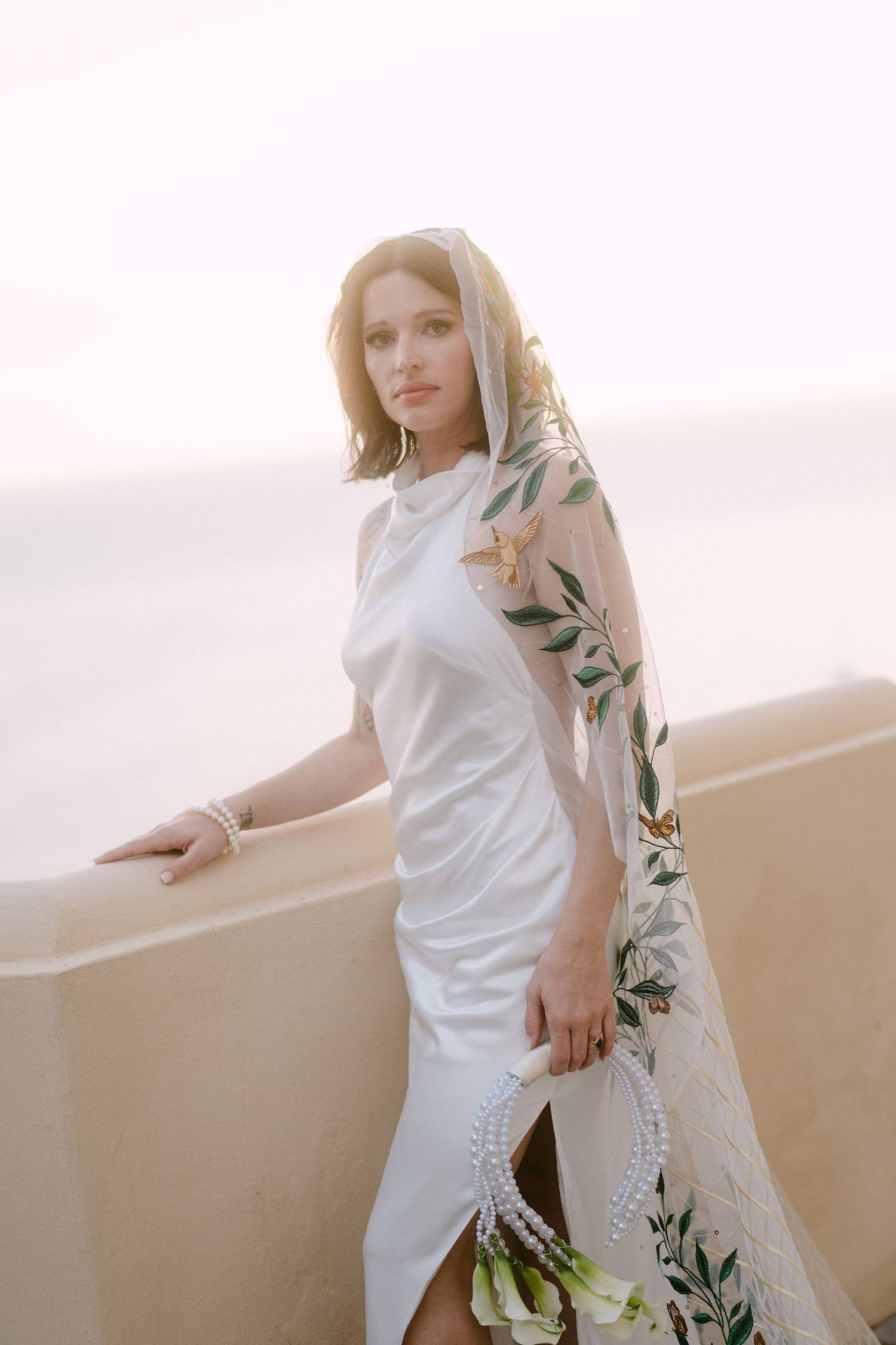 A person in white attire poses against a light backdrop with delicate botanical elements.