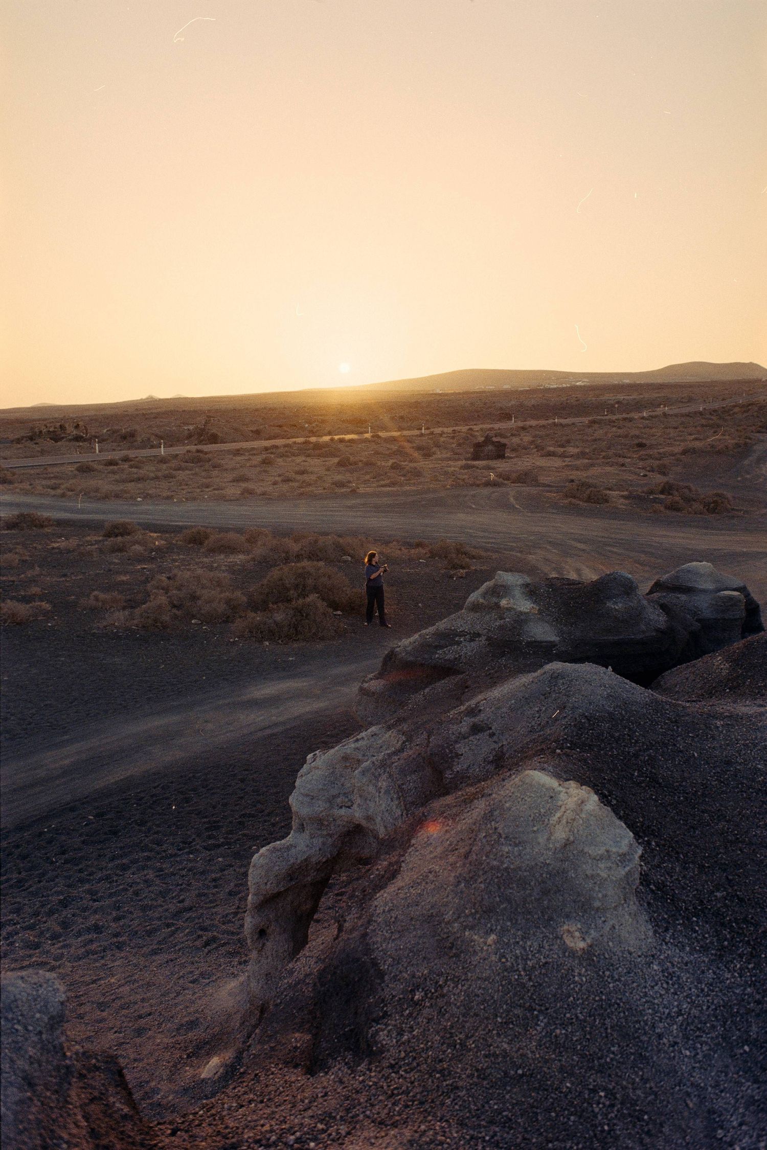 A small figure stands on desert terrain at sunset with rocky outcrops and mountains silhouetted against an orange sky.