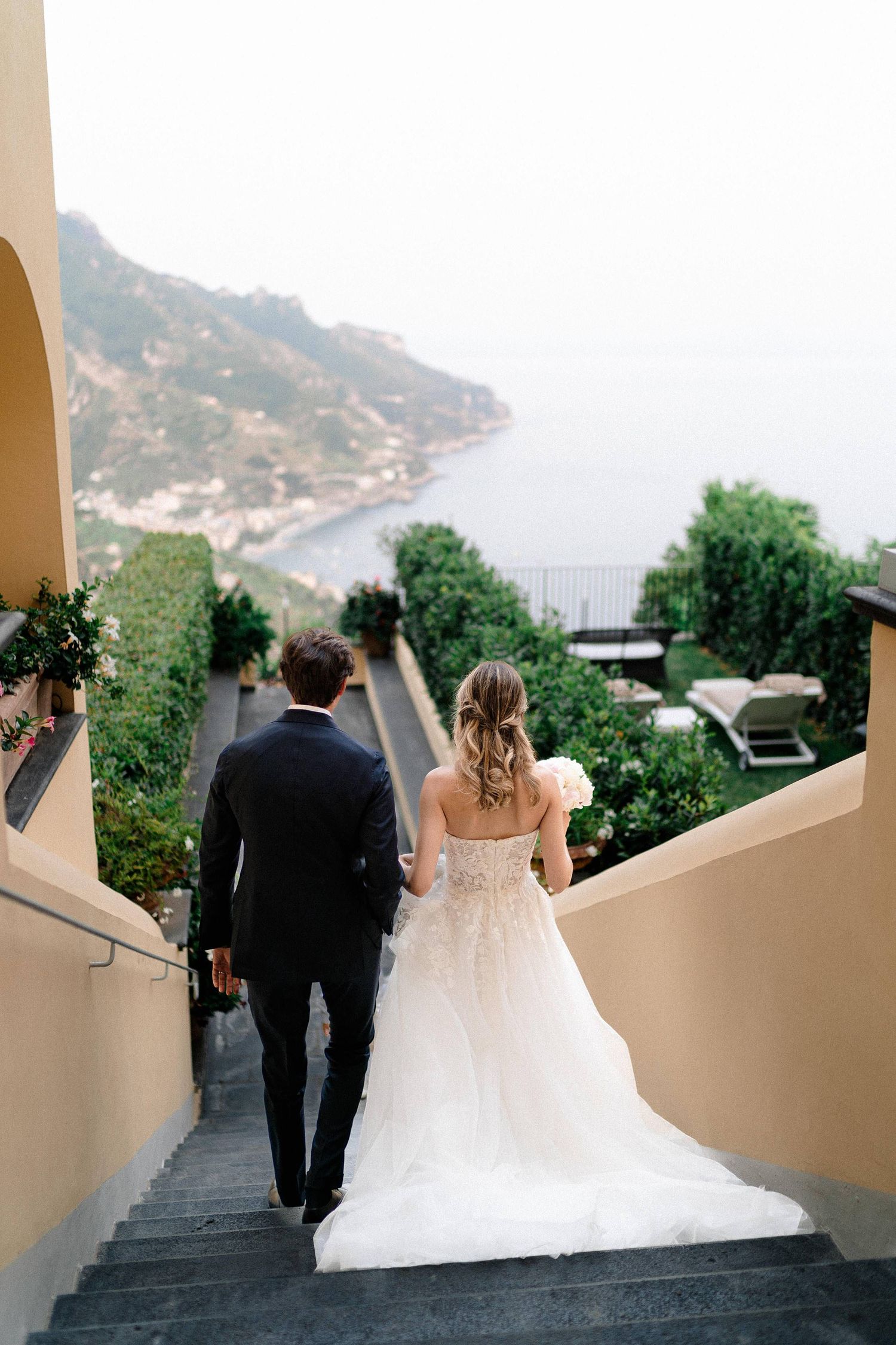 A couple descends stone steps overlooking a scenic coastal view in their wedding attire.