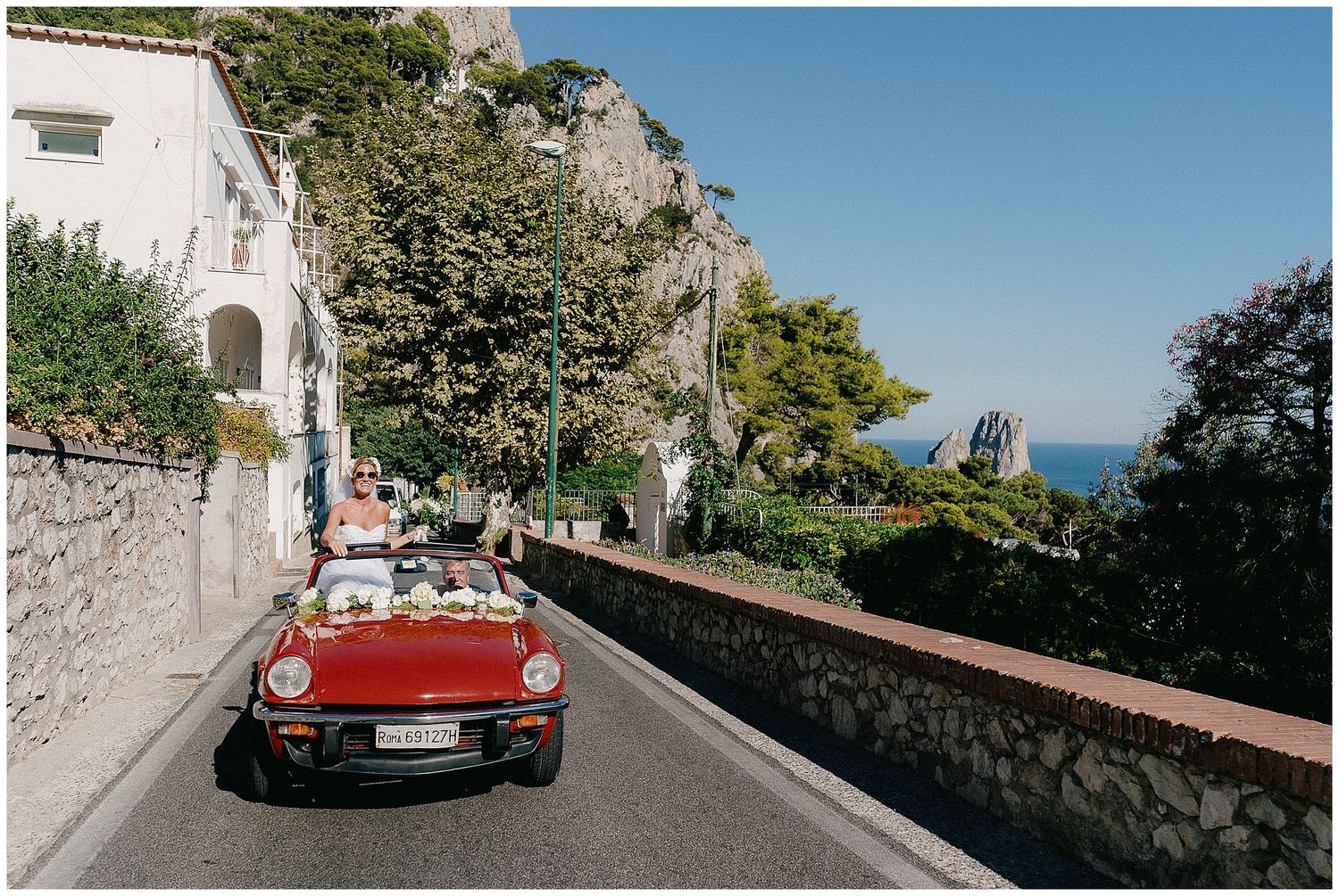 A red vintage convertible drives along a scenic coastal road in Capri with cliffside views and Mediterranean vegetation.