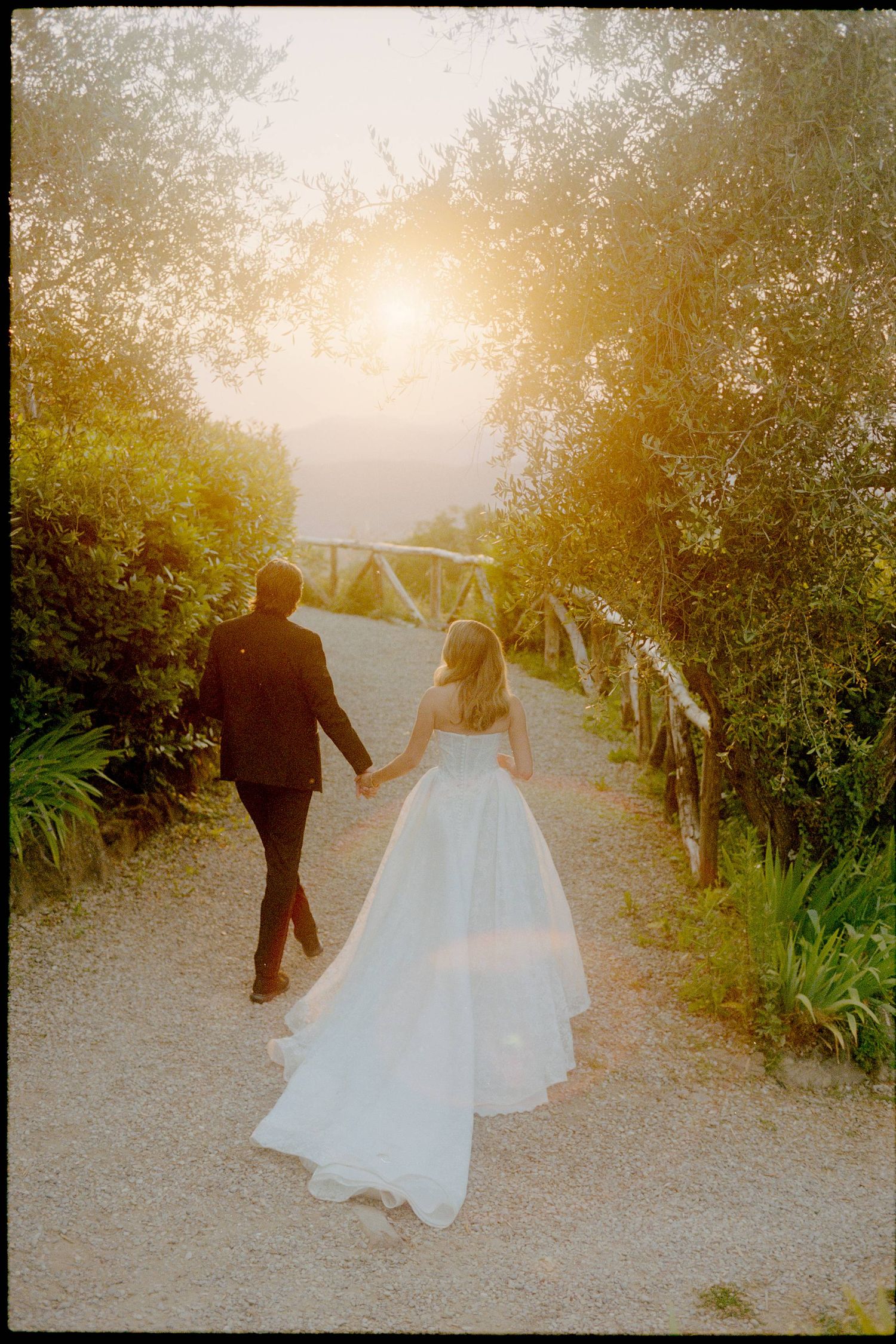 Bride and groom walking hand in hand down a sunlit garden path, bride in flowing white gown, golden hour light.