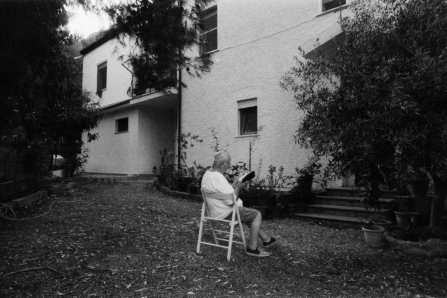 A figure sits alone in a white folding chair on a grassy yard beside a white building in black and white photography.