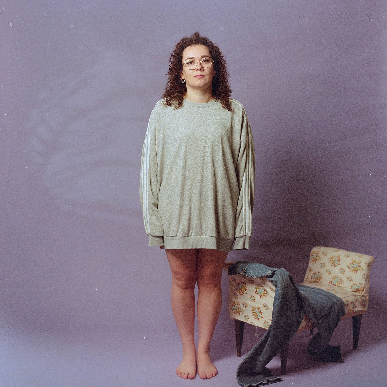Person wearing an oversized beige sweater stands barefoot against purple background with a broken chair nearby.