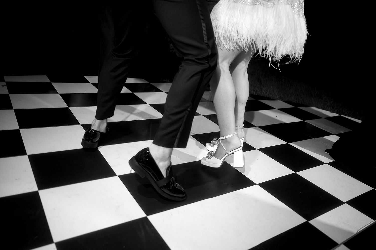 Black and white image of two pairs of feet dancing on a checkered floor.