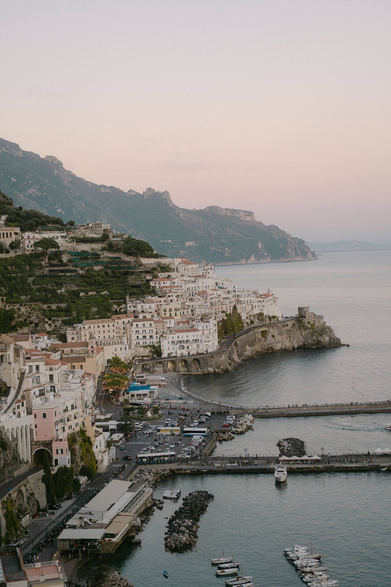 Scenic aerial view of Amalfi Coast in Italy showing white buildings cascading down cliffs and boats in harbor at sunset.