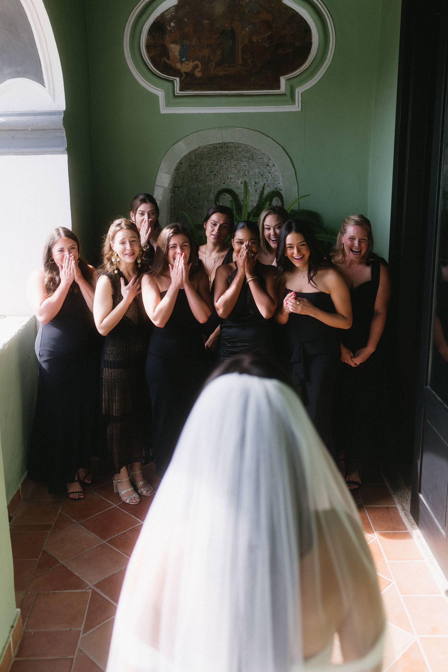 A bride in a white gown stands with bridesmaids wearing black dresses covering their faces in a playful photo pose.
