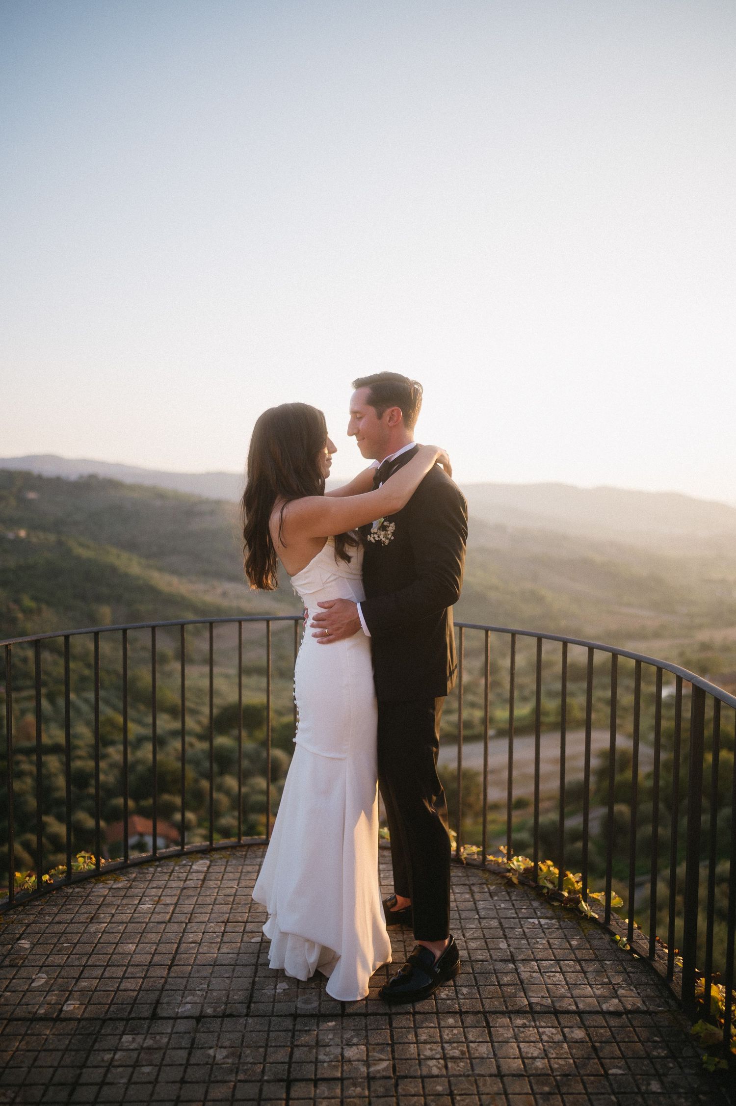 A couple embraces on a scenic overlook at sunset with rolling hills in the background.