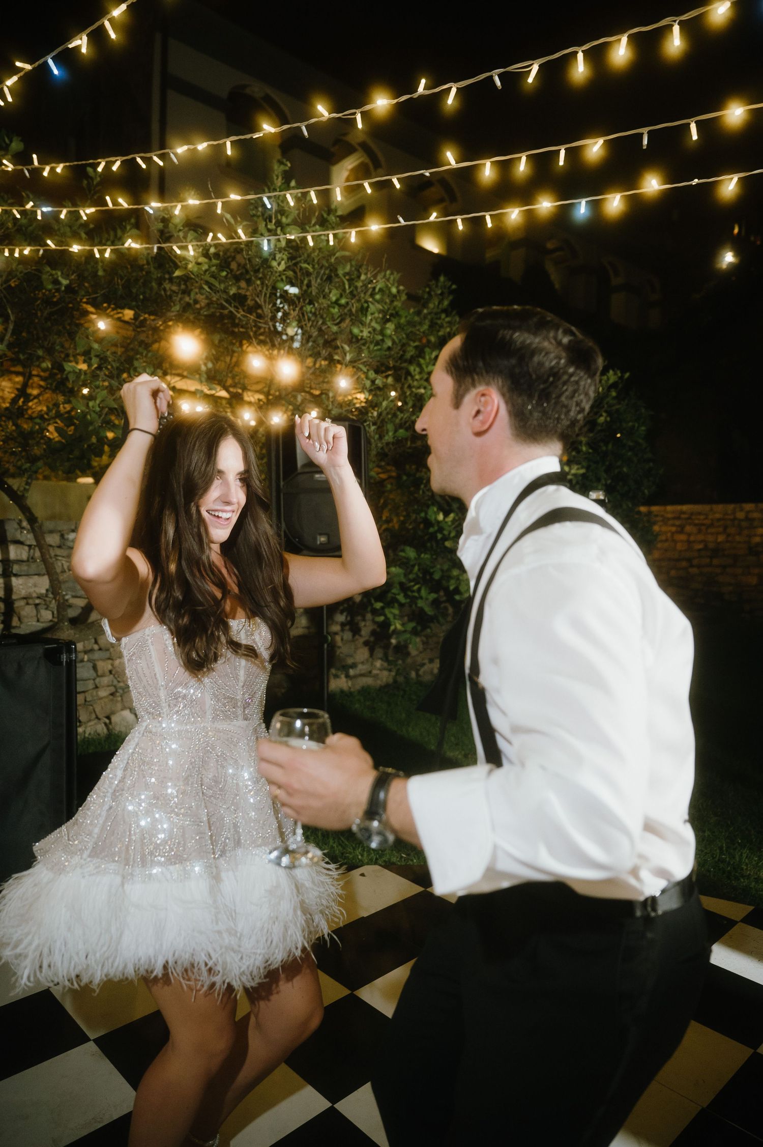 A couple dances together under glowing string lights at an outdoor evening celebration.