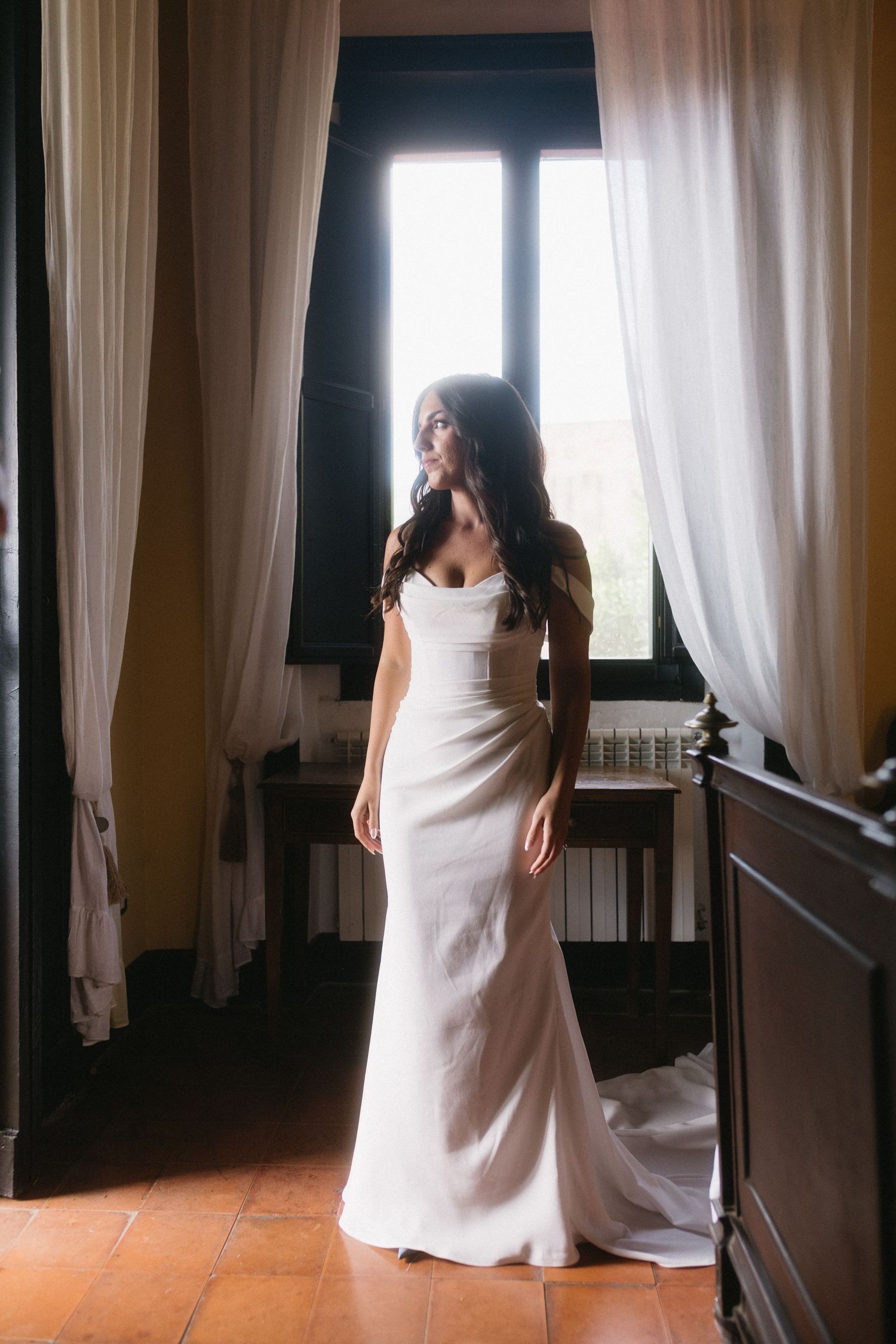 A figure in a white dress stands by billowing sheer curtains in a dimly lit room with wooden floors.