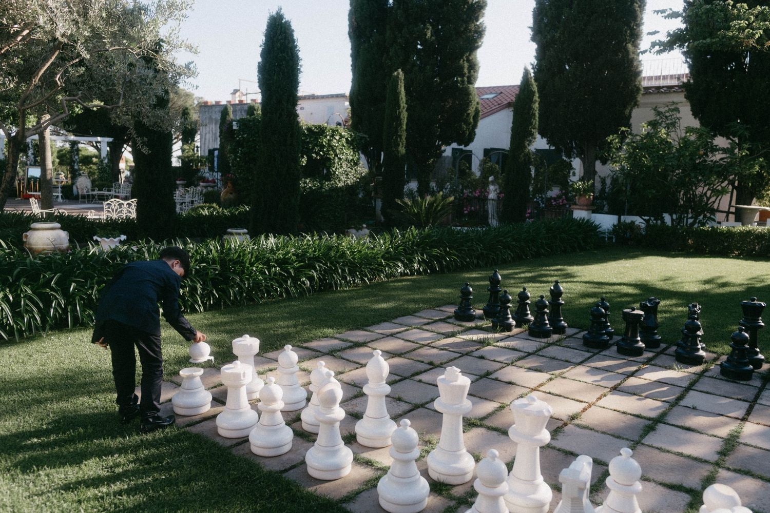 A giant outdoor chess set with white and black pieces stands on a paved surface in a garden setting.