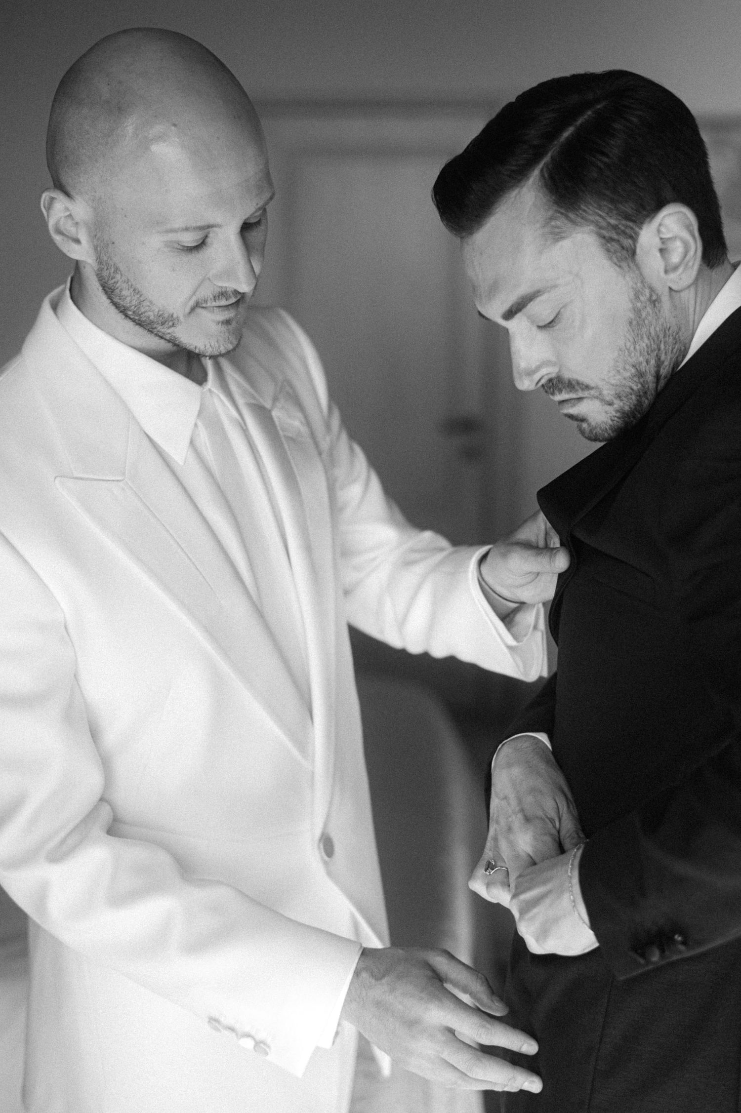 Two men in formal attire prepare for a special occasion, one helping the other adjust clothing in black and white.