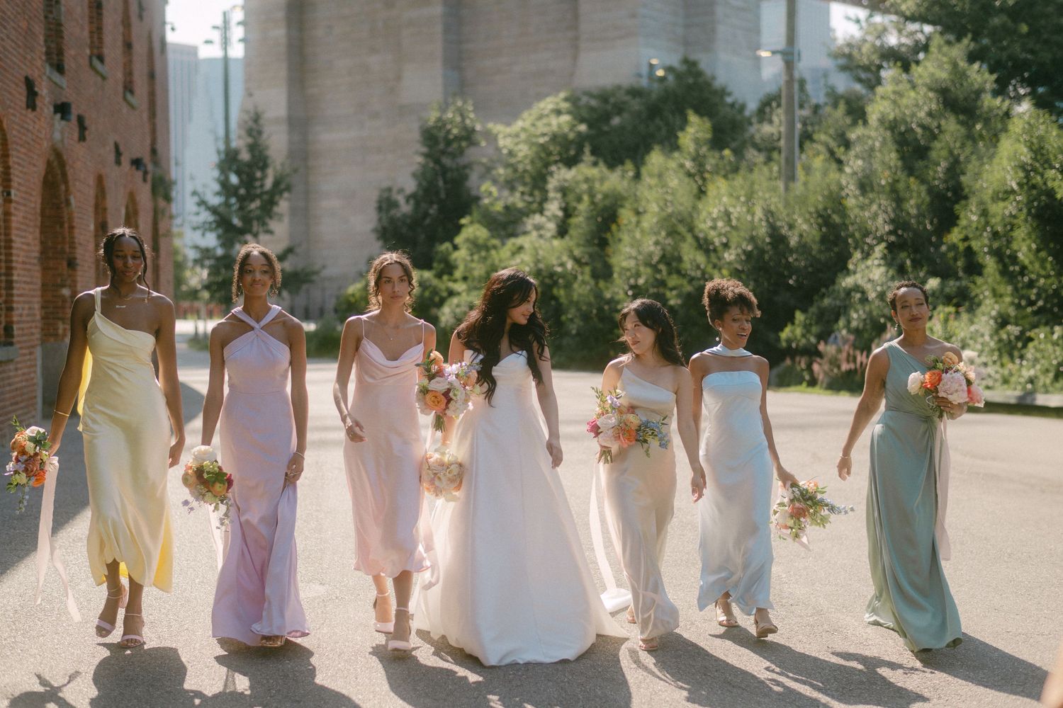 Bridesmaids in pastel dresses stand in line against a stone wall at an outdoor wedding venue.