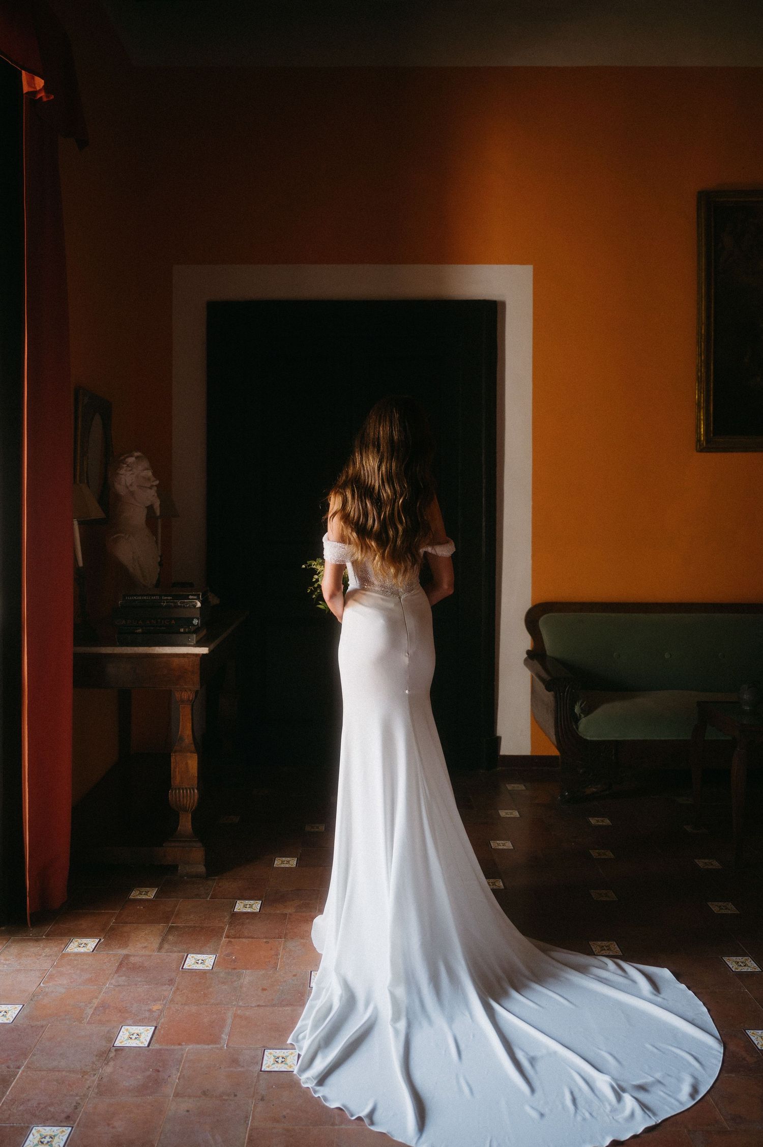 A bride in a flowing white wedding gown walks across a rustic terracotta floor in a dimly lit room.