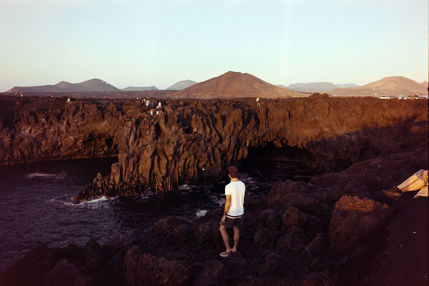 A person stands on rugged cliffs overlooking crashing waves against rocky coastline at dusk with mountains in the distance.