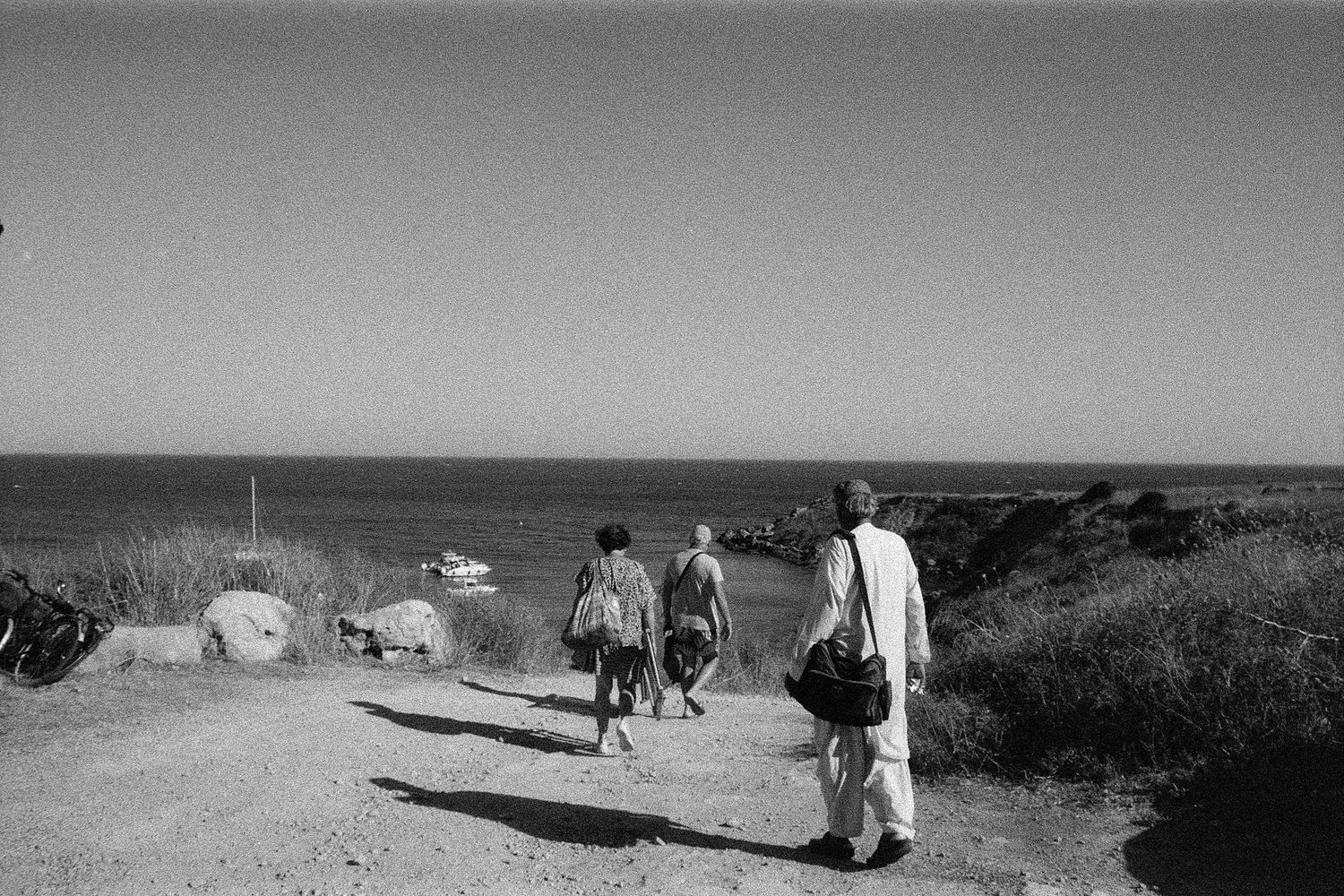 People walking along a coastal path towards parked boats in a black and white seaside landscape.