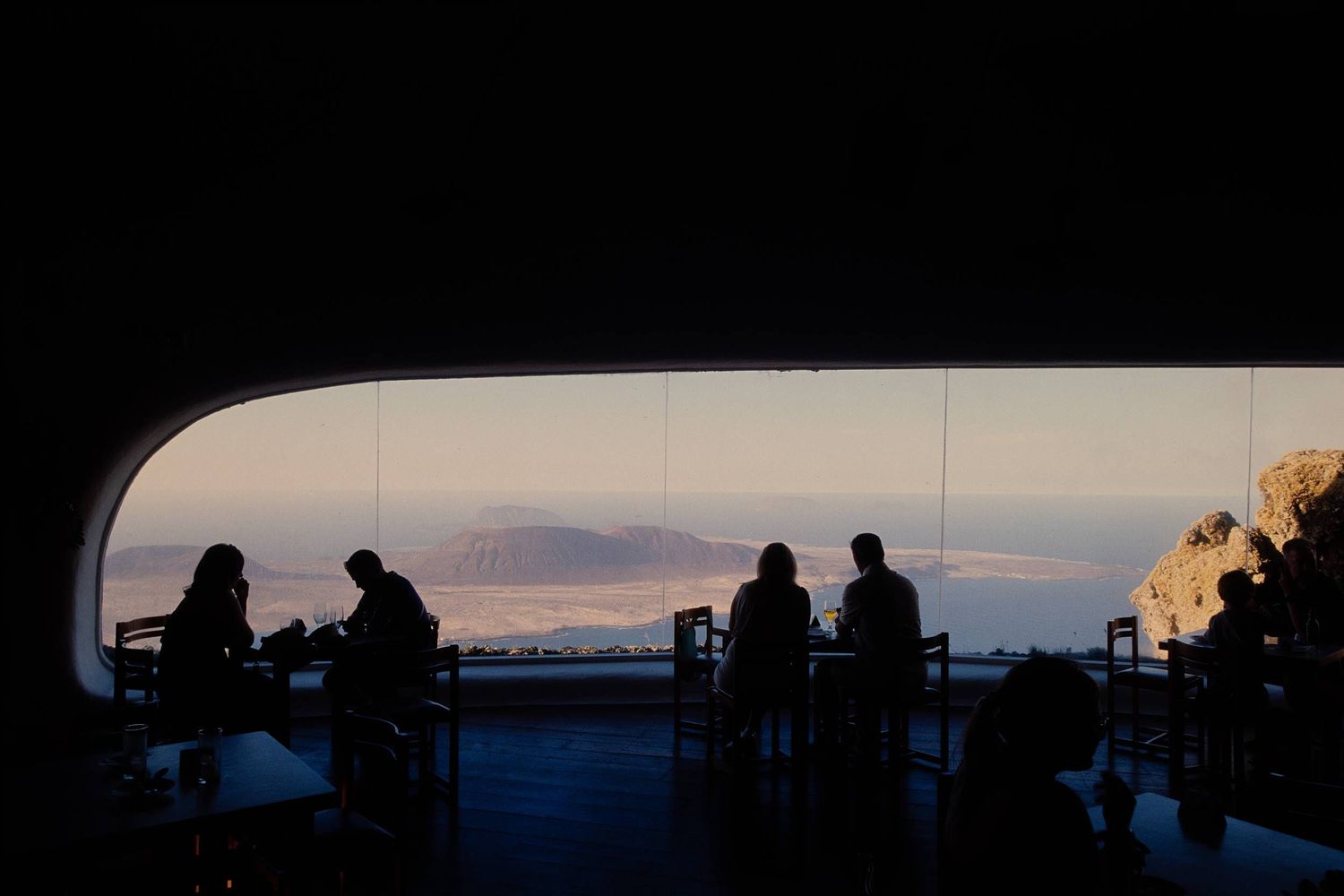 Silhouetted figures gather under an archway viewing a scenic sunset over distant mountains and water.