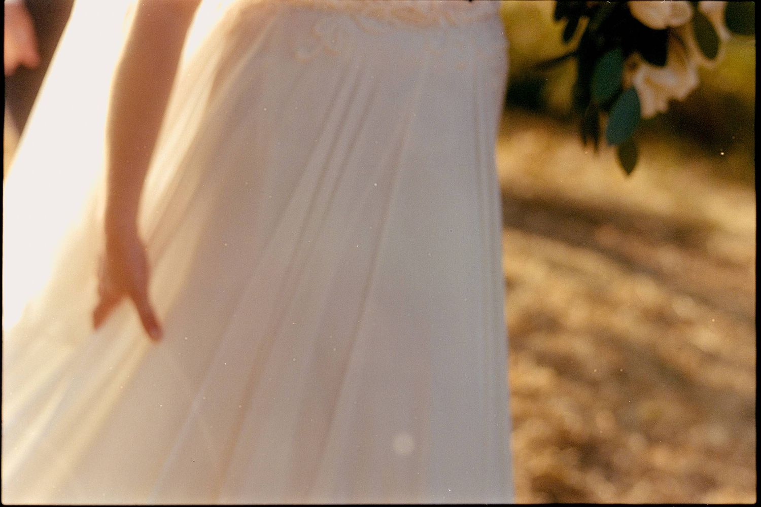 A soft, flowing white wedding dress fabric swaying in sunlit outdoor setting with blurred foliage in background.