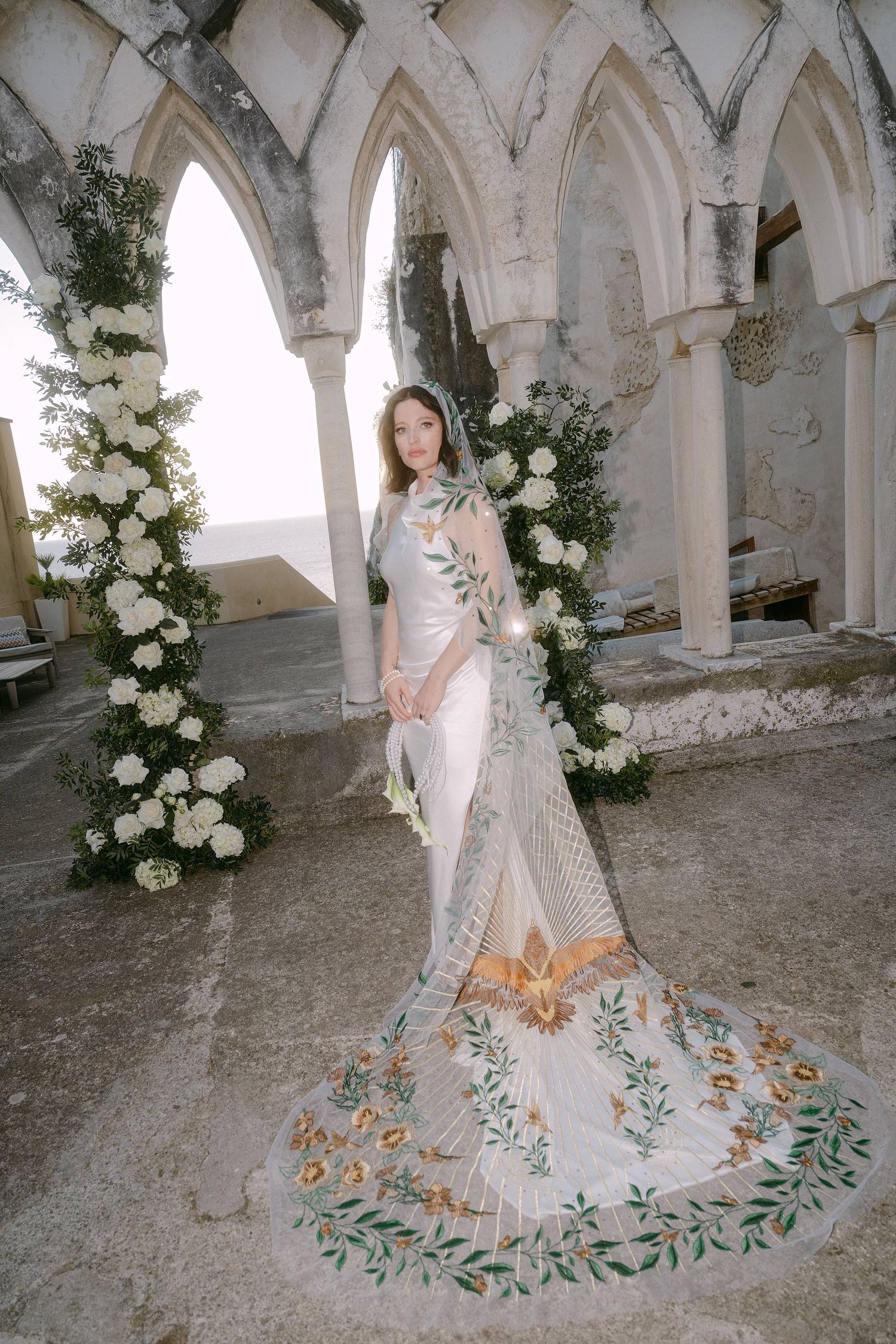 A bridal photoshoot in an ornate stone archway featuring a flowing white gown with intricate floral embroidered train details.