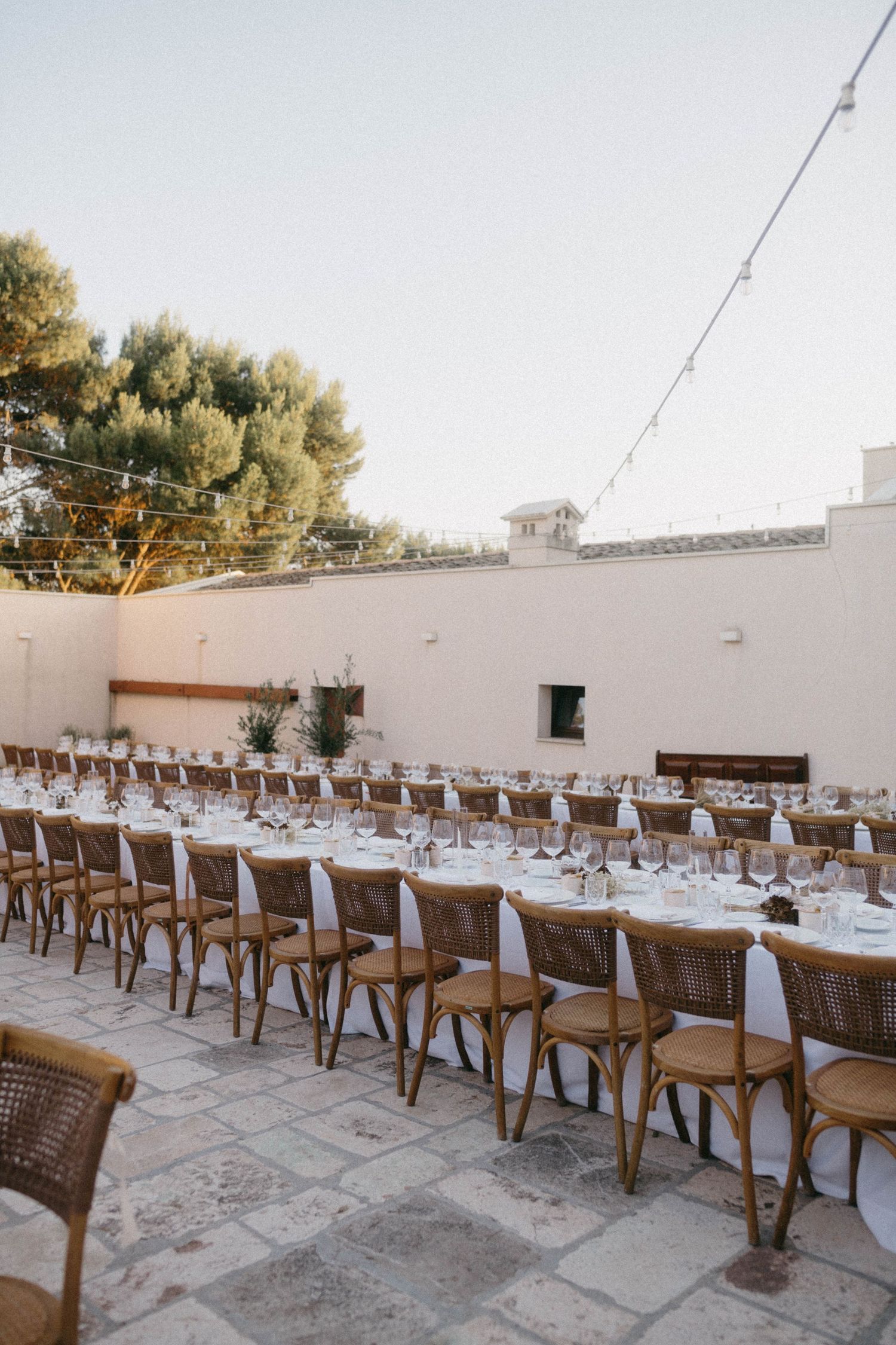 Long rows of wooden chairs and white-clothed tables set up for an outdoor wedding reception on a stone patio.