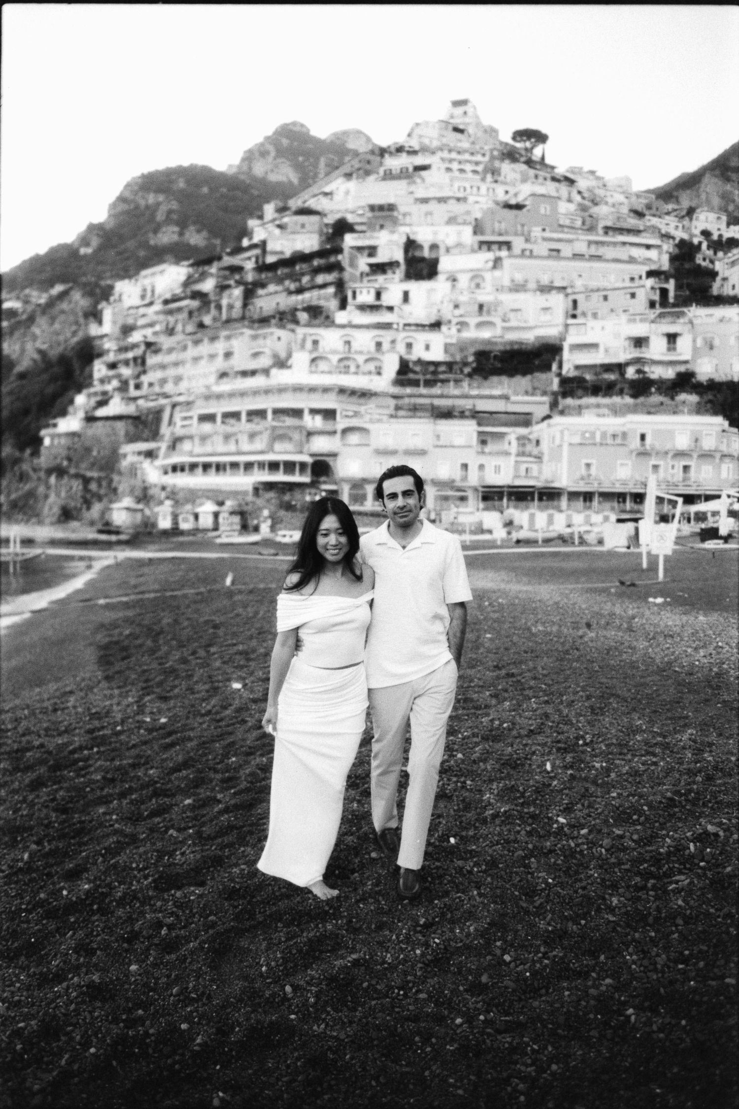 Vintage black and white photos show a couple posing on a beach with hillside Mediterranean buildings in background.