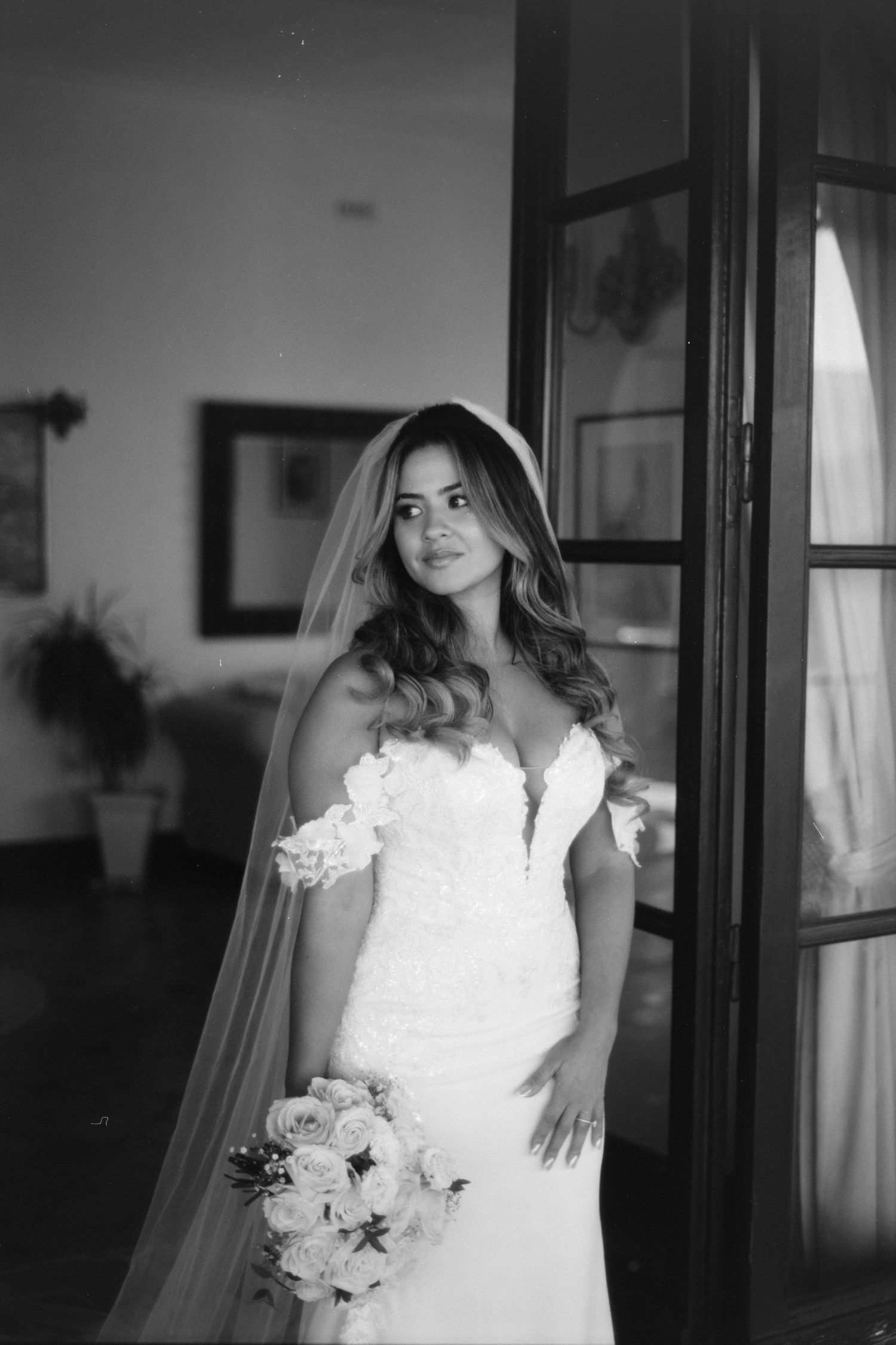 A bride in an elegant white wedding dress poses by a window in a series of black and white portraits.