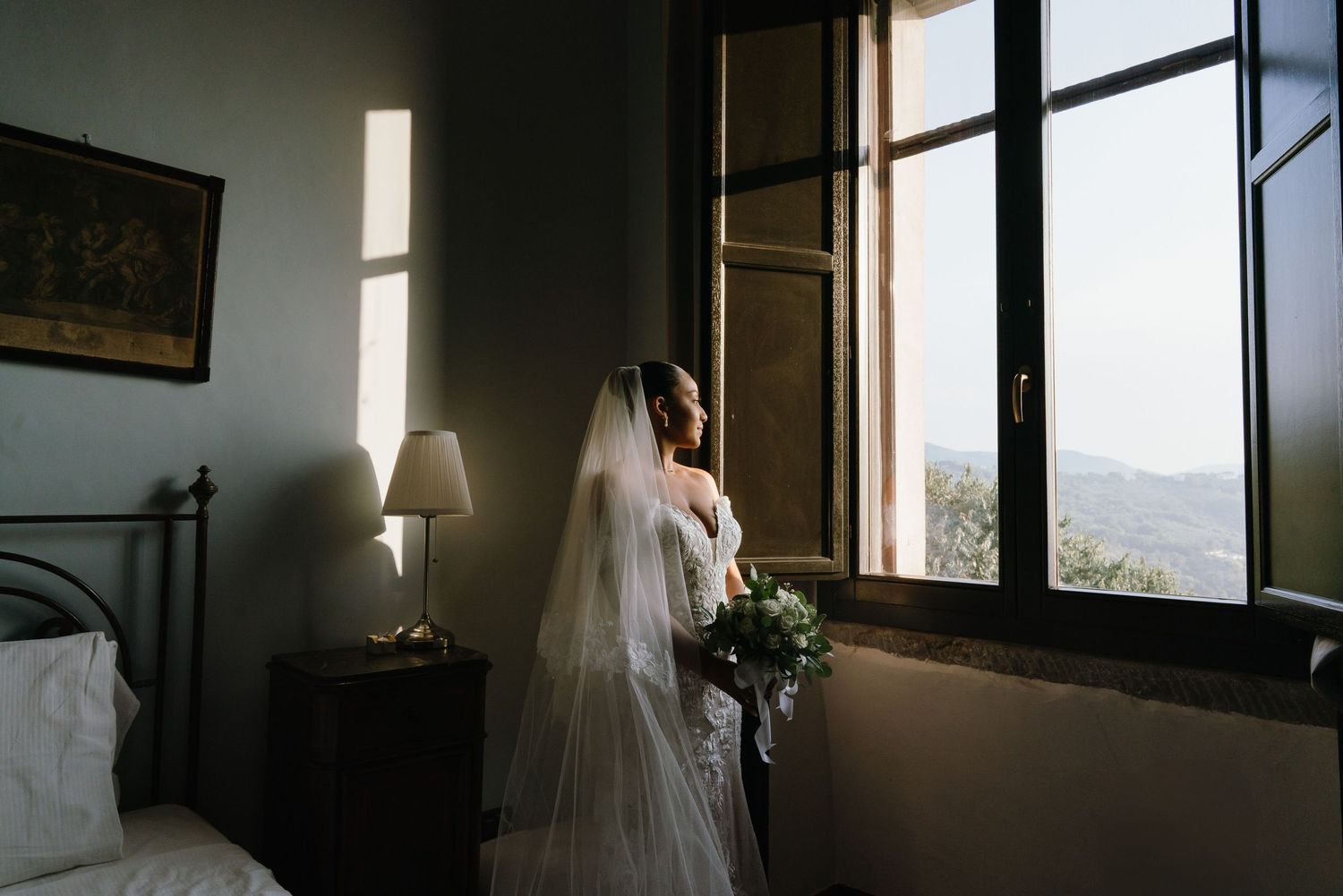 A bride in a white wedding dress stands by a bright window in a rustic interior space.