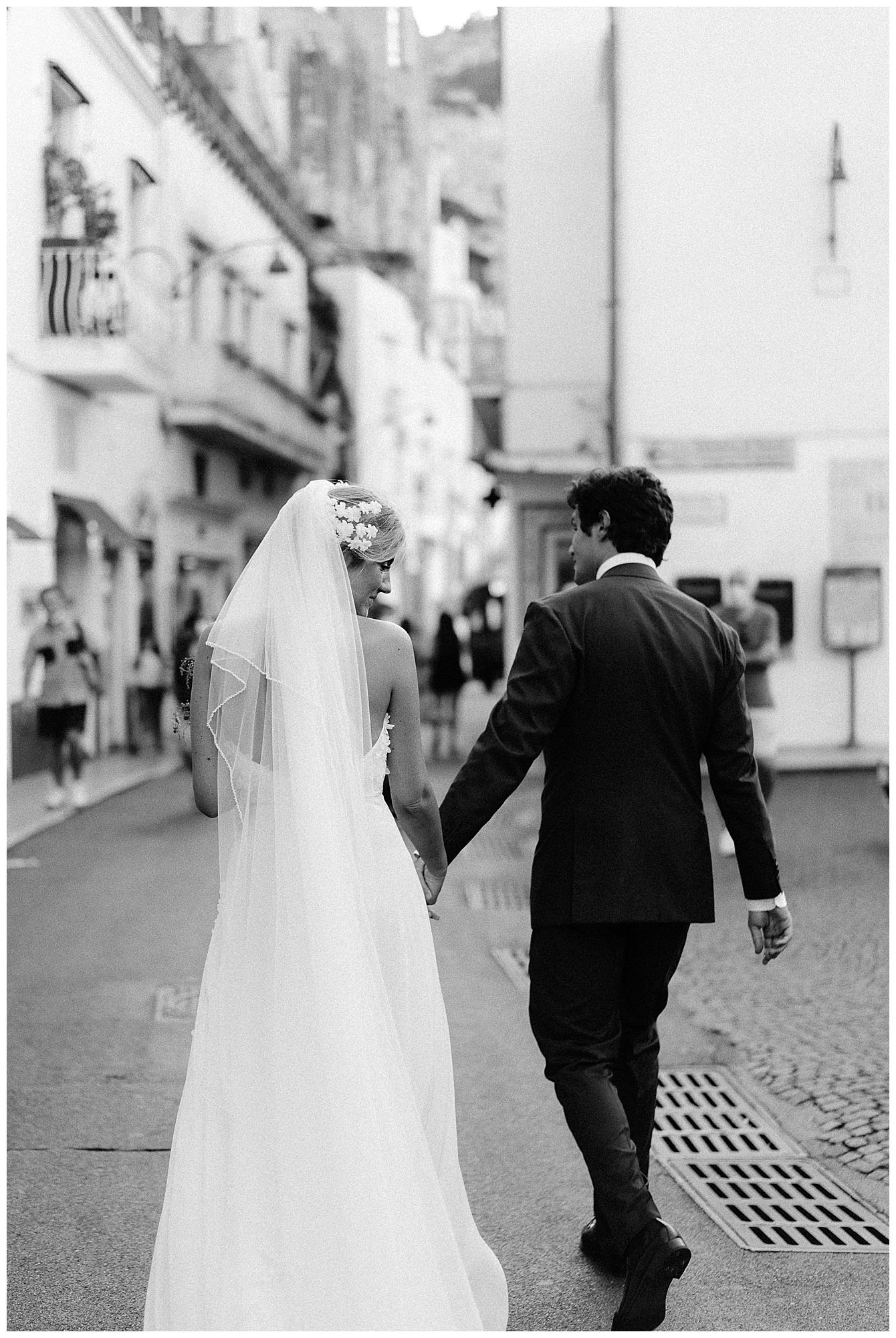 A bride and groom walking down a cobblestone street in a black and white photo.