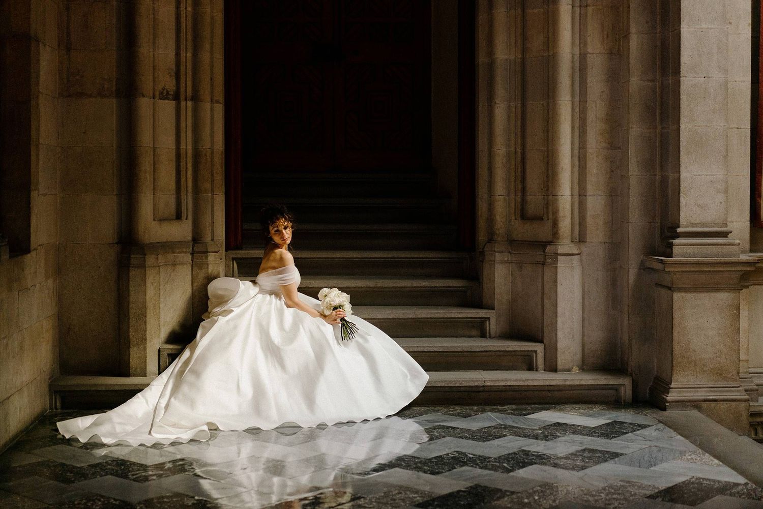 A bride in a voluminous white wedding gown sits elegantly on stone steps between classical columns in dramatic lighting.
