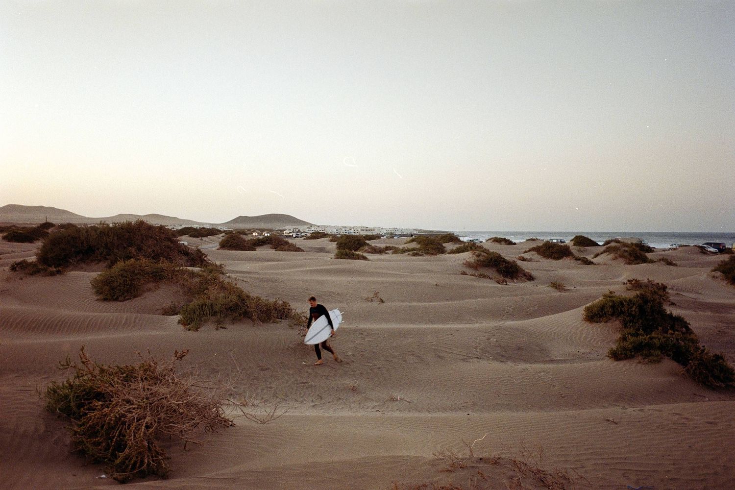 A lone figure carrying a surfboard walks across sandy dunes at dusk with ocean in the background.