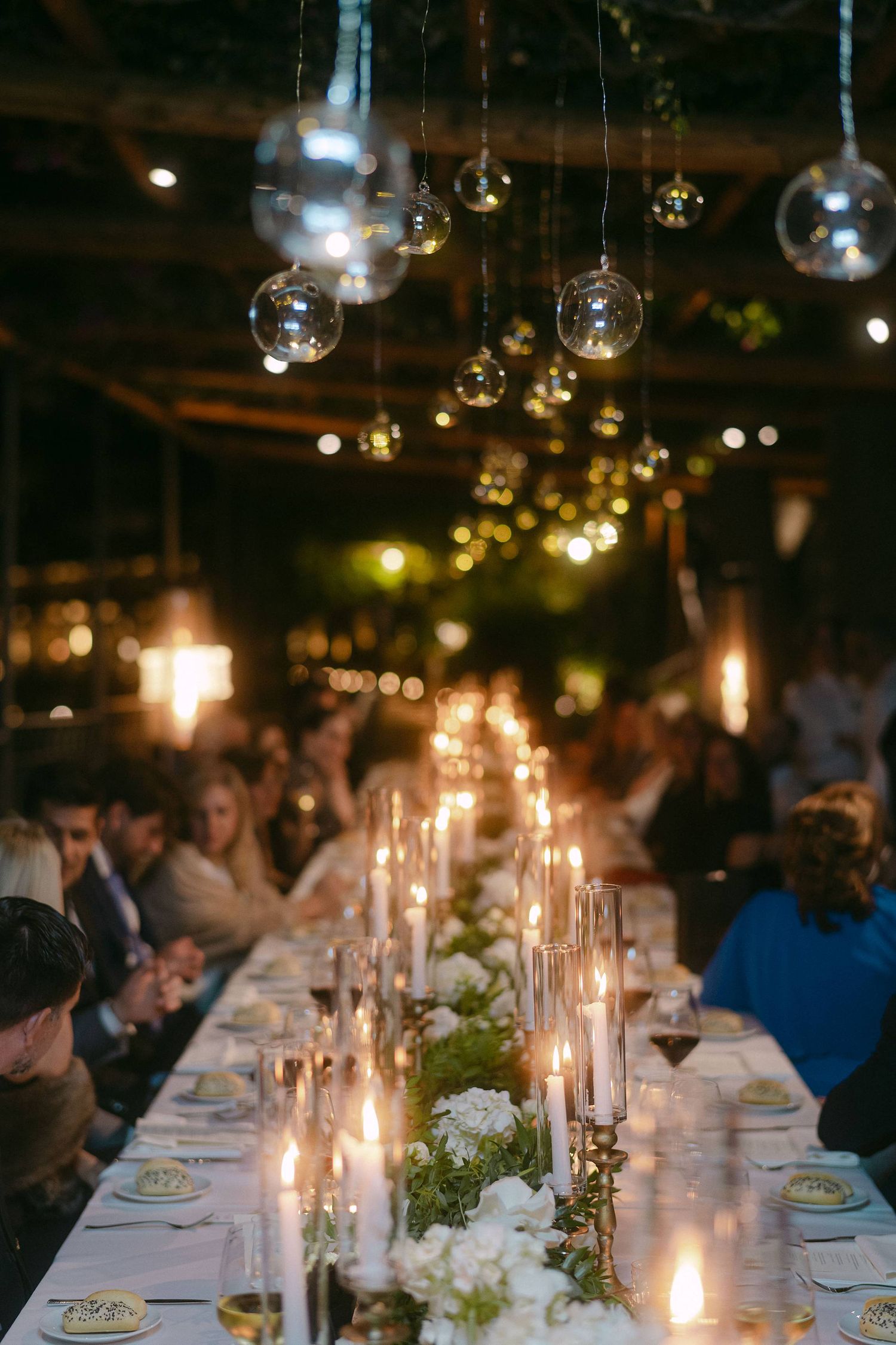 Long wedding reception table decorated with white flowers and glowing candles under hanging glass orb lights creates romantic ambiance.