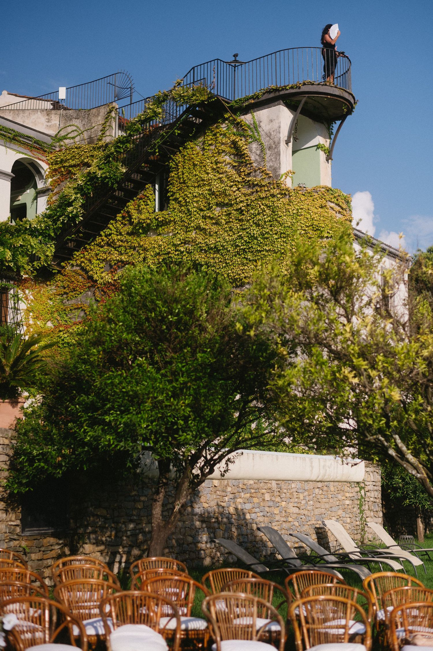 Rustic outdoor dining area with wooden chairs arranged on a stone patio beneath ivy-covered walls and blue sky.