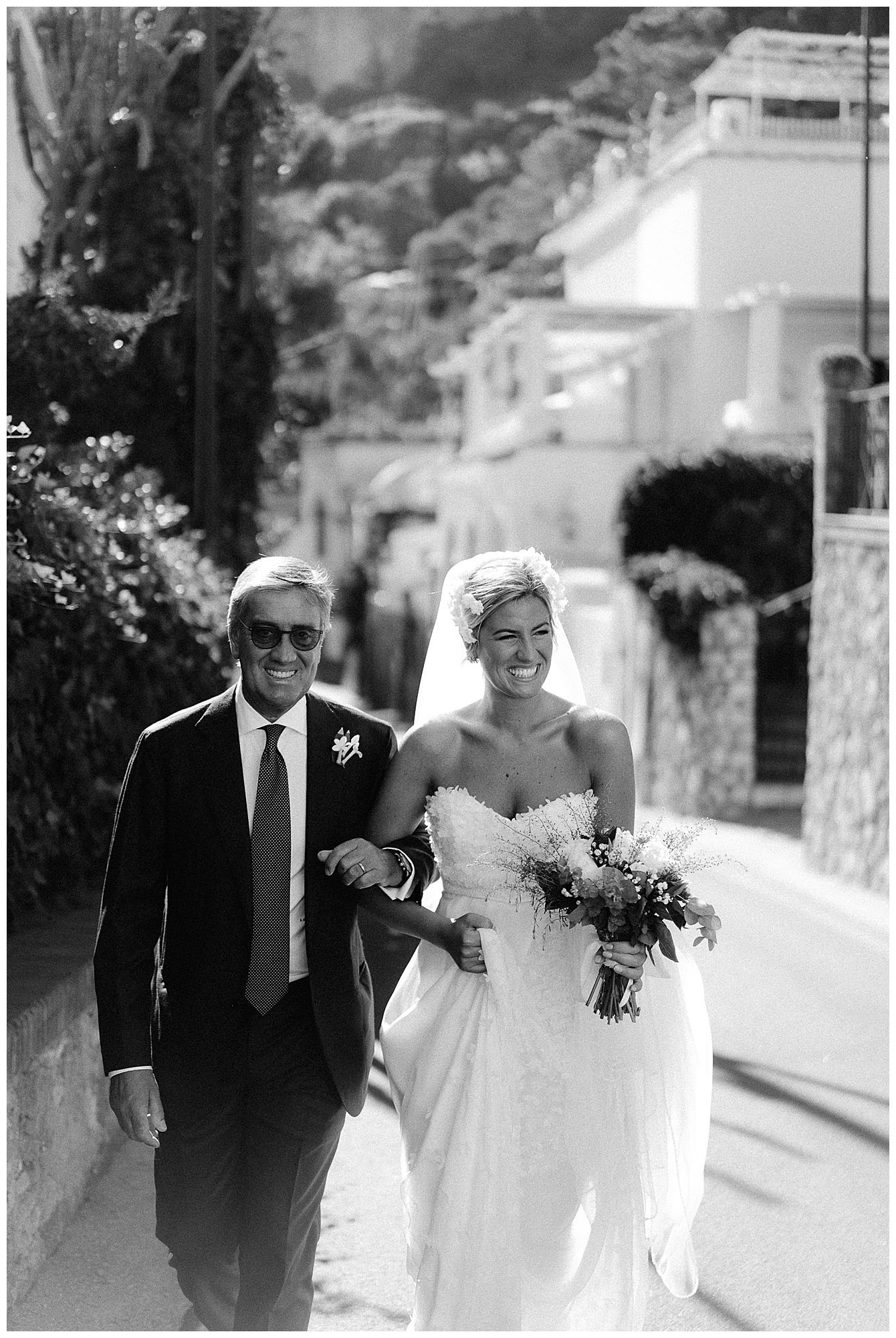 A woman in a white wedding dress walks arm in arm with a man in a dark suit on a sunny Mediterranean street.
