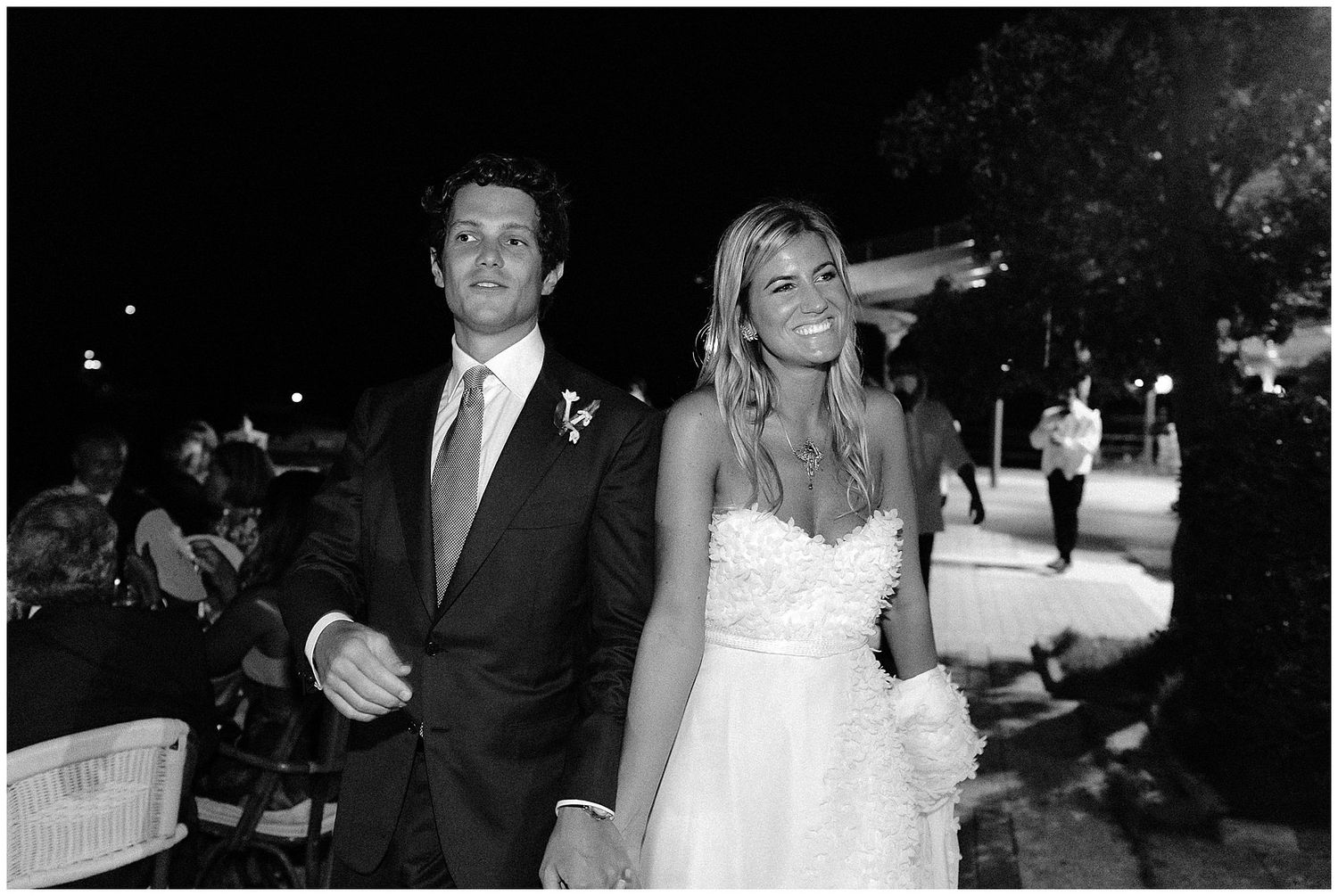 A black and white photograph of a couple walking outdoors at night, with the woman wearing a white dress.