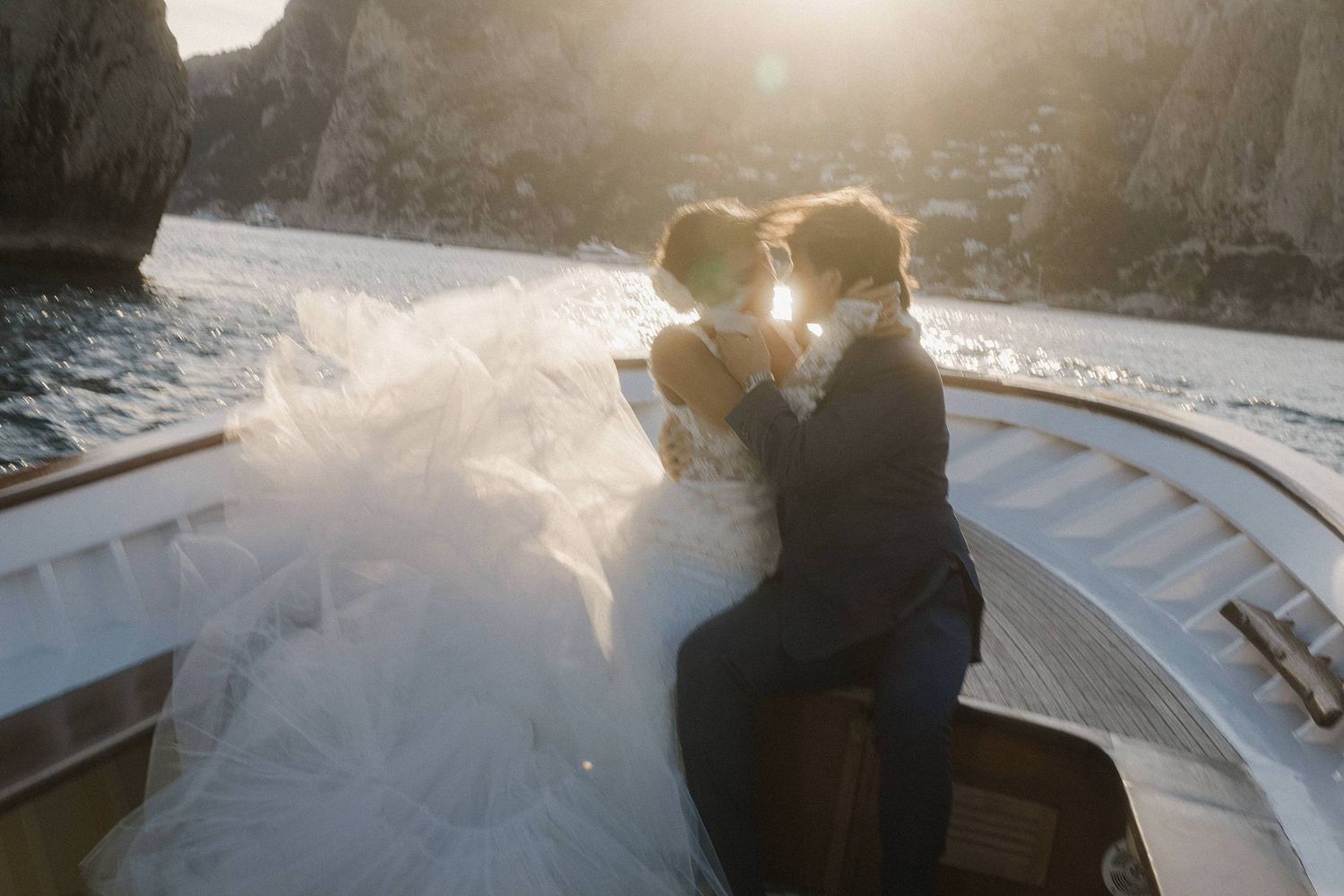 Bride and groom share a romantic kiss on a boat at sunset with stunning coastal cliffs in the background.