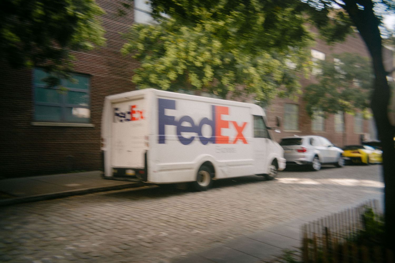 A FedEx delivery van parked on a residential street with brick buildings and trees in the background.
