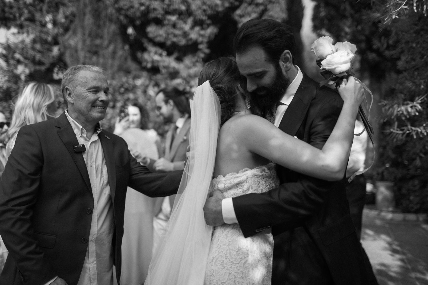 Black and white photo of a wedding ceremony moment showing a couple kissing while others watch in an outdoor garden setting.