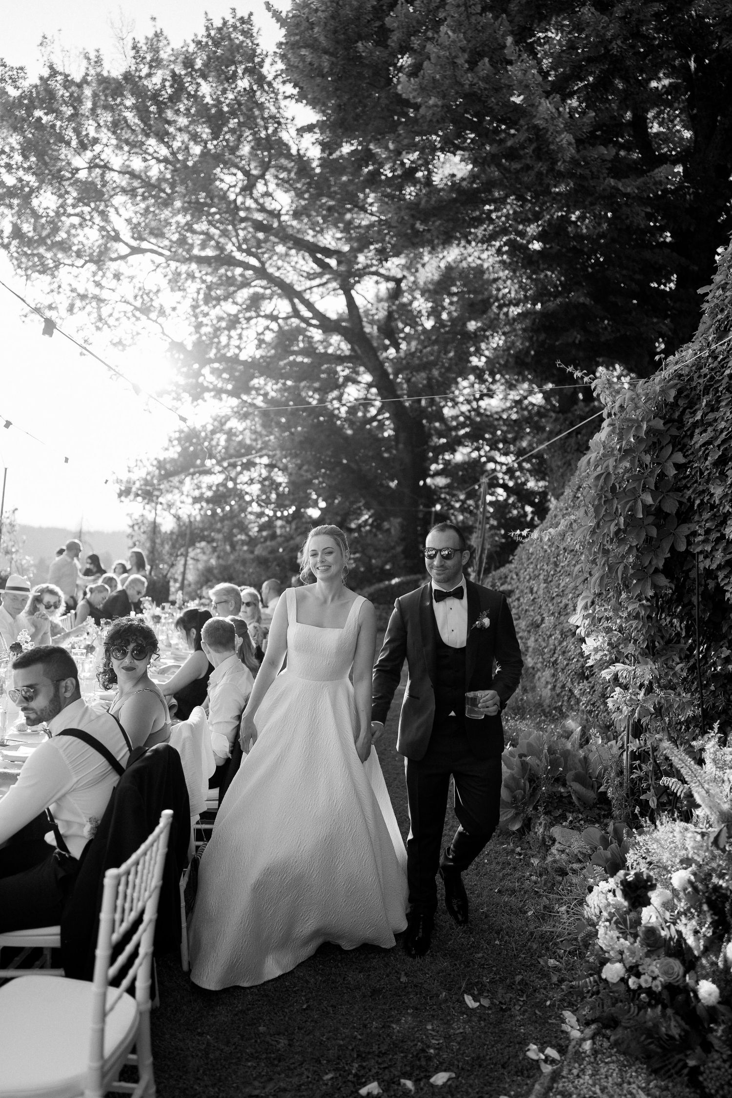 A wedding party dances and celebrates outdoors under tree cover during a joyful reception celebration.