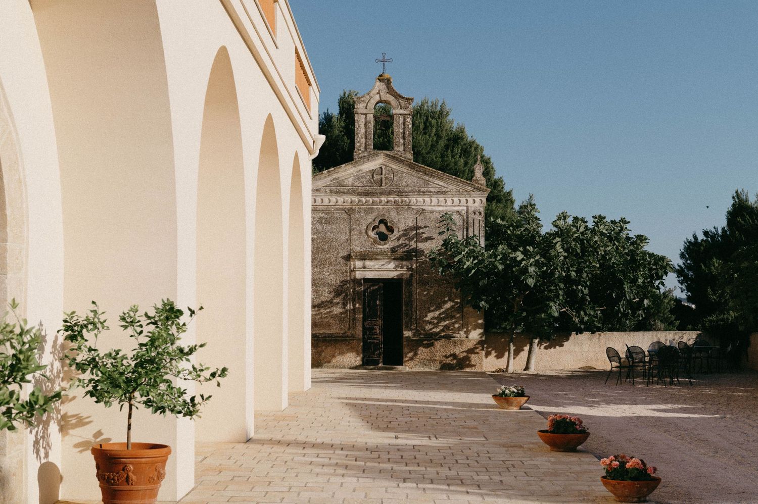 Small stone chapel entrance with arched doorway and bell tower against white building walls and potted plants in Mediterranean setting.