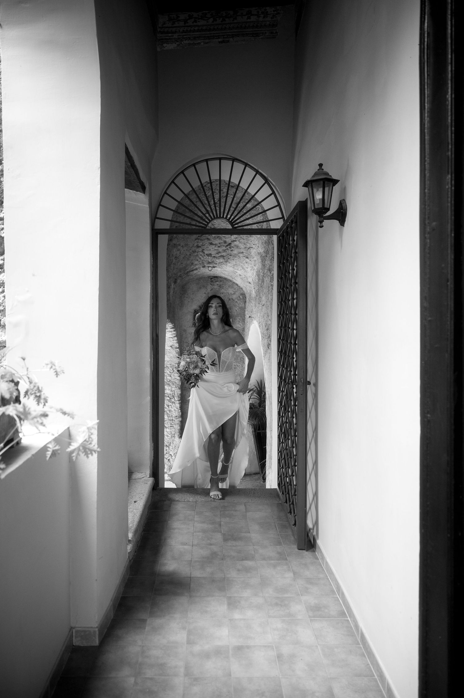Black and white photograph of a figure in a white dress standing in an elegant hallway with an arched doorway and wall lantern.