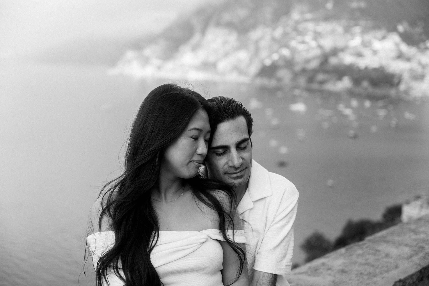 A romantic black and white photo of a couple embracing on a cliff overlooking the ocean on a cloudy day.