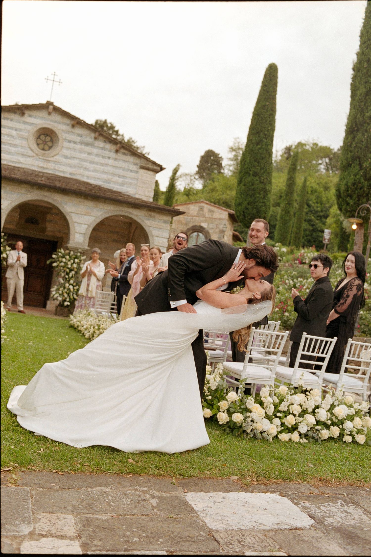 Bride and groom share a romantic dip kiss at outdoor wedding ceremony with white floral arrangements and stone chapel.