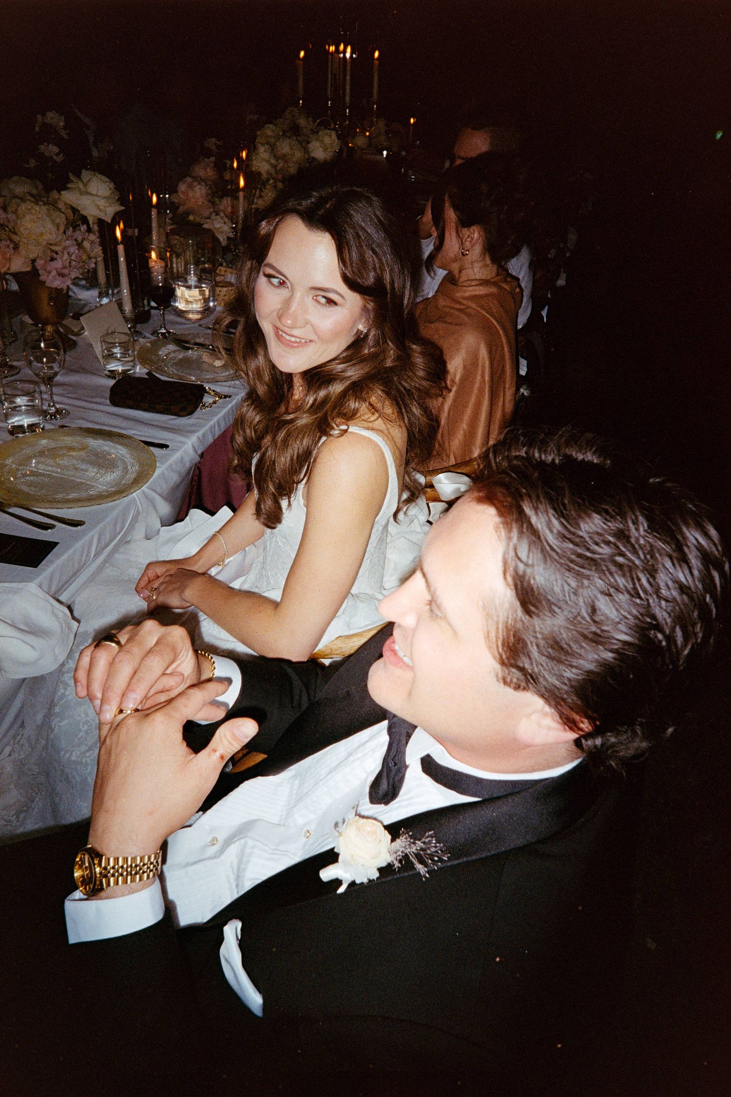 Elegant couple at formal dinner, woman in light dress smiling, man in tuxedo, candlelit table setting.