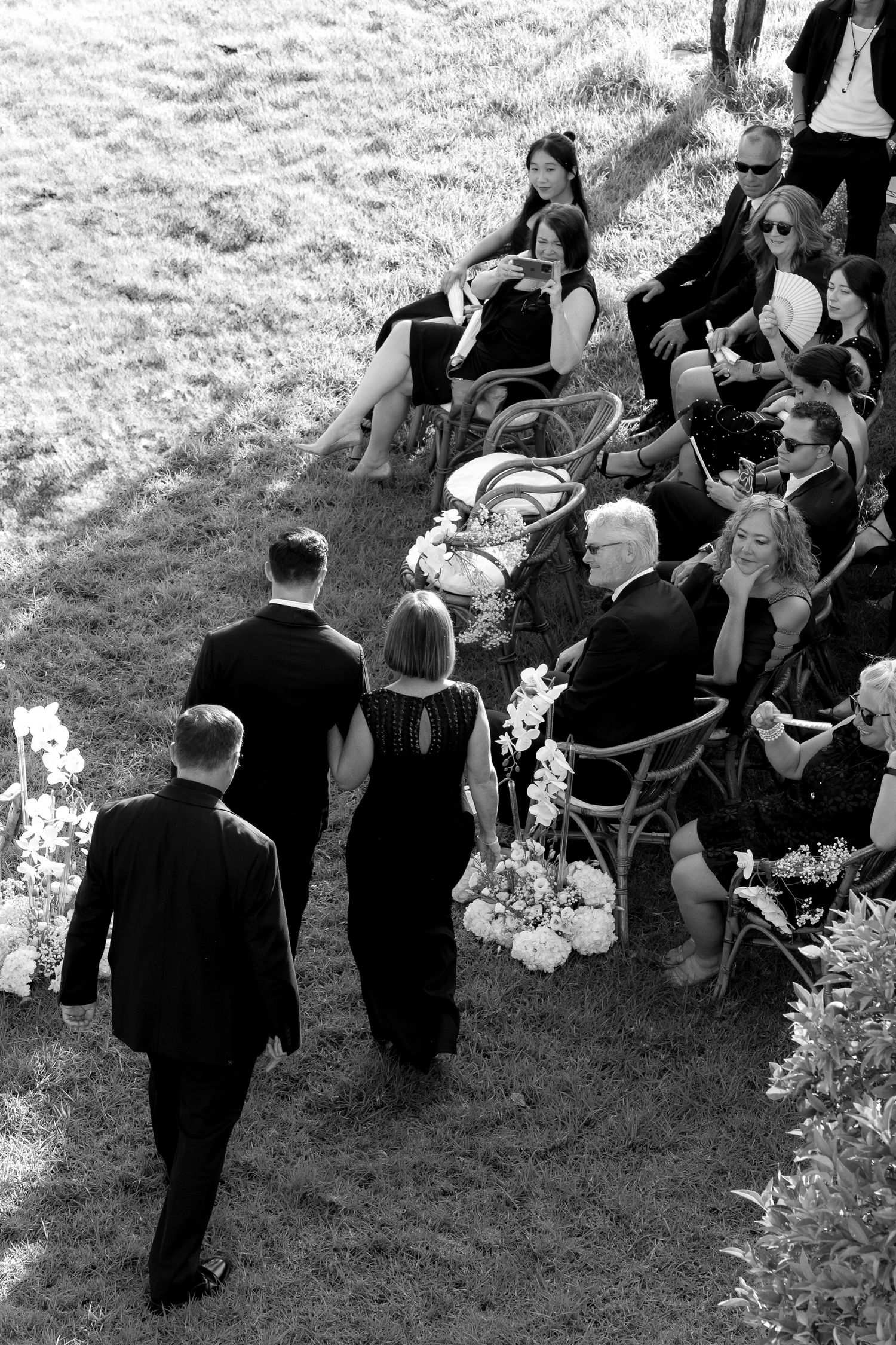 Aerial view of a wedding ceremony with guests seated outdoors in black attire surrounded by floral arrangements.