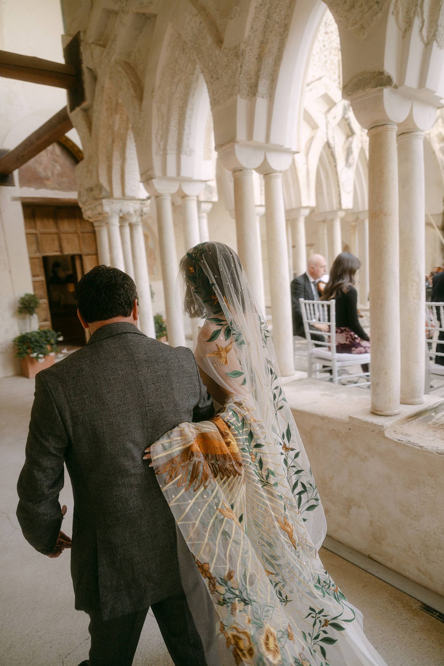 A bride in an embroidered veil walks with her companion through an elegant columned archway at a wedding venue.
