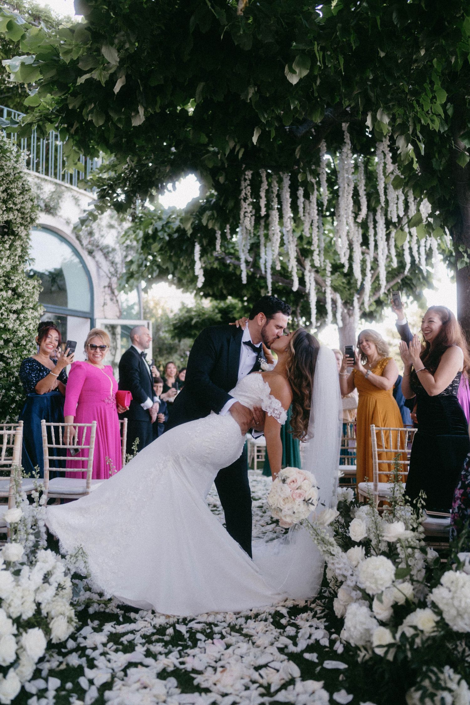 Romantic outdoor wedding ceremony with white floral arrangements and hanging crystals under a tree canopy.