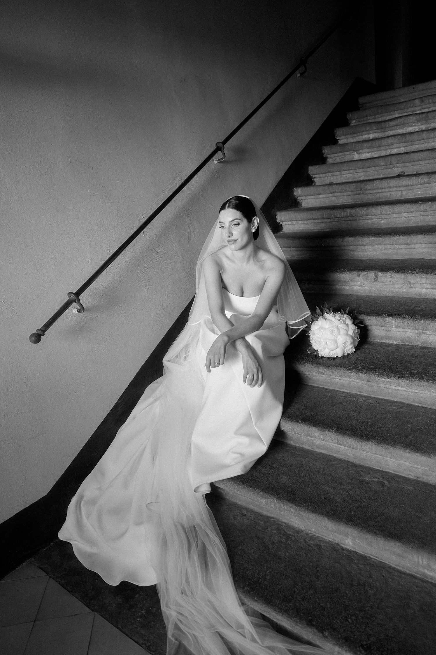 Bride in white gown and veil sits on stone staircase holding bouquet, elegant black and white wedding portrait.