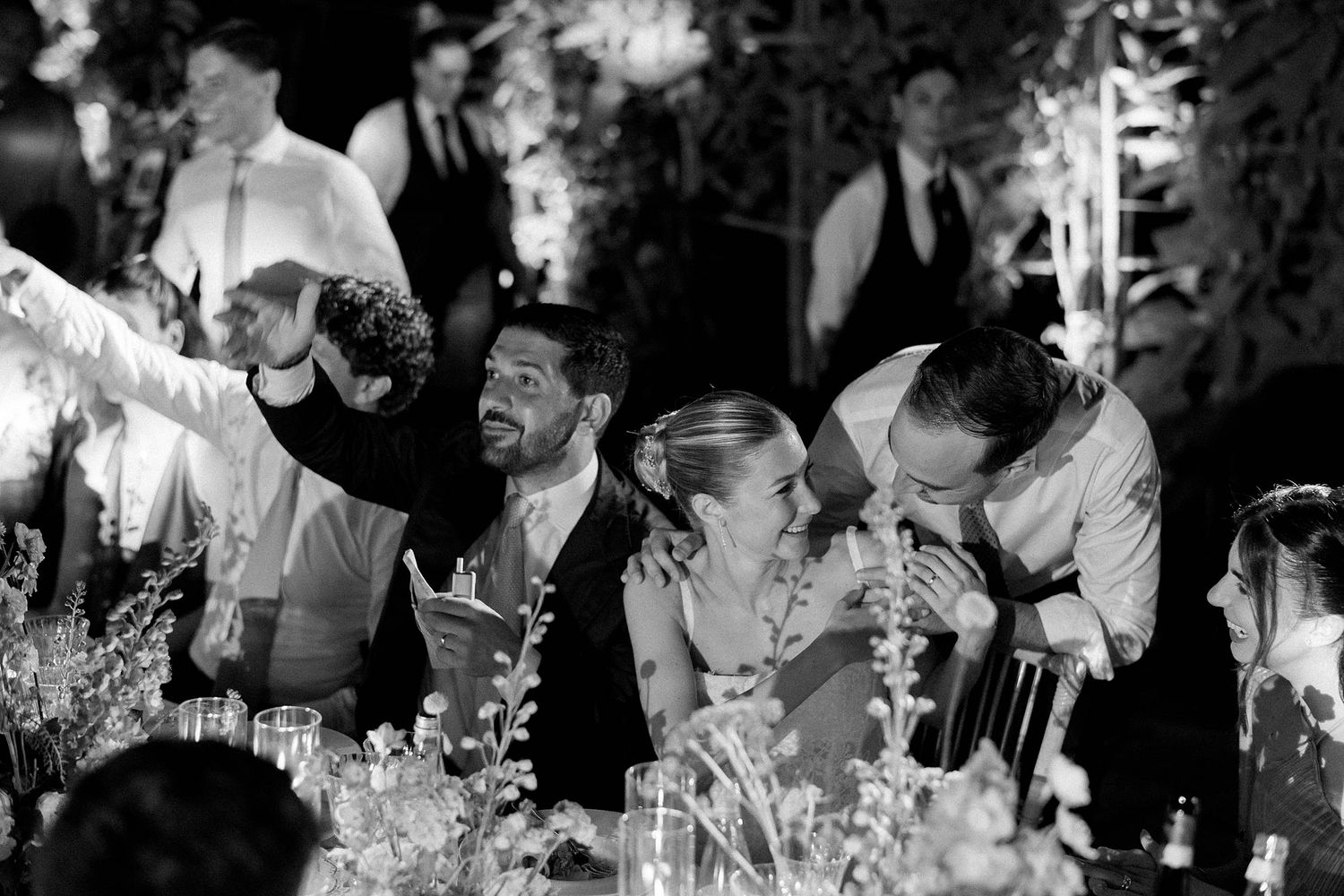 Black and white wedding reception photo of guests laughing and celebrating at an elegantly decorated outdoor dinner table.