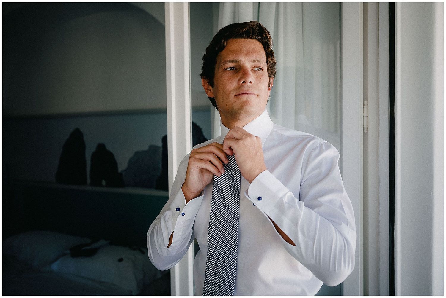 Person adjusting tie while getting dressed in crisp white shirt by window.