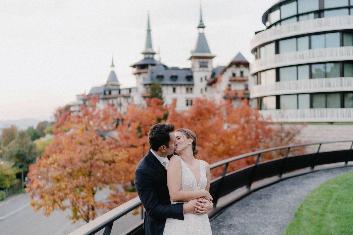 A couple embraces outdoors on an autumn day with dramatic castle-like architecture and orange foliage in the background.