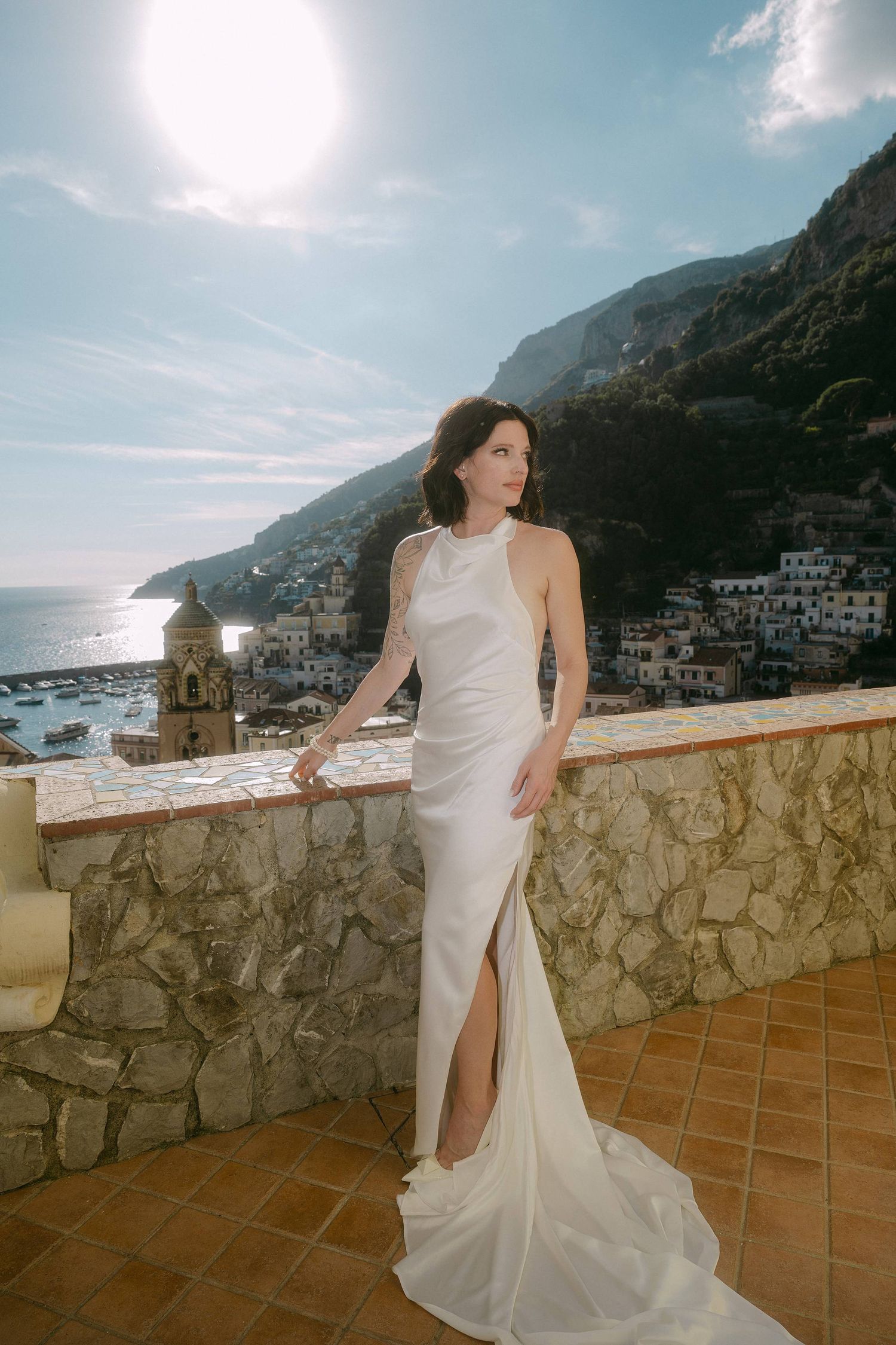 A person in a flowing white dress poses on a terrace overlooking the scenic Amalfi Coast in Italy during sunset.